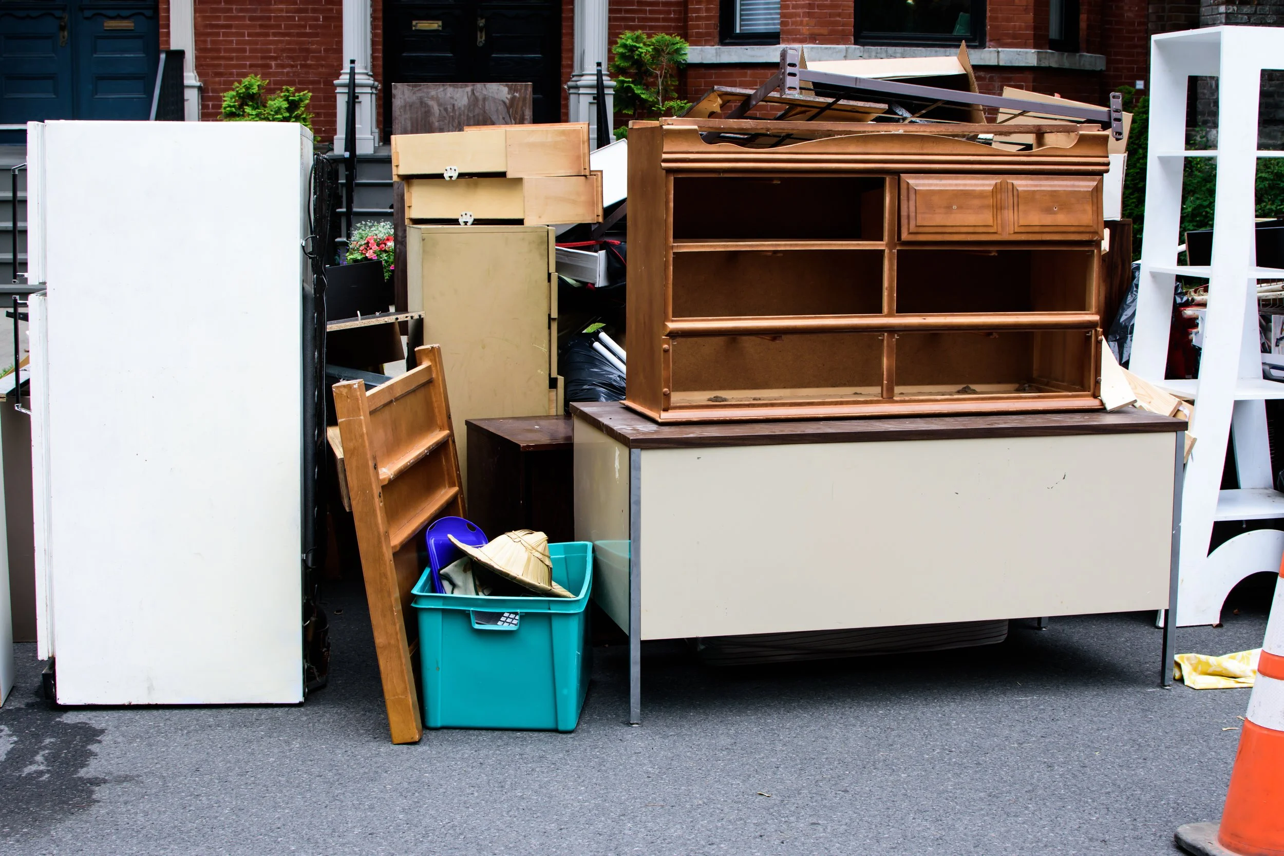 A collection of furniture and household items, including a wooden dresser, white cabinets, a small table, and various other furniture pieces, placed outdoors on a street in front of a brick building.