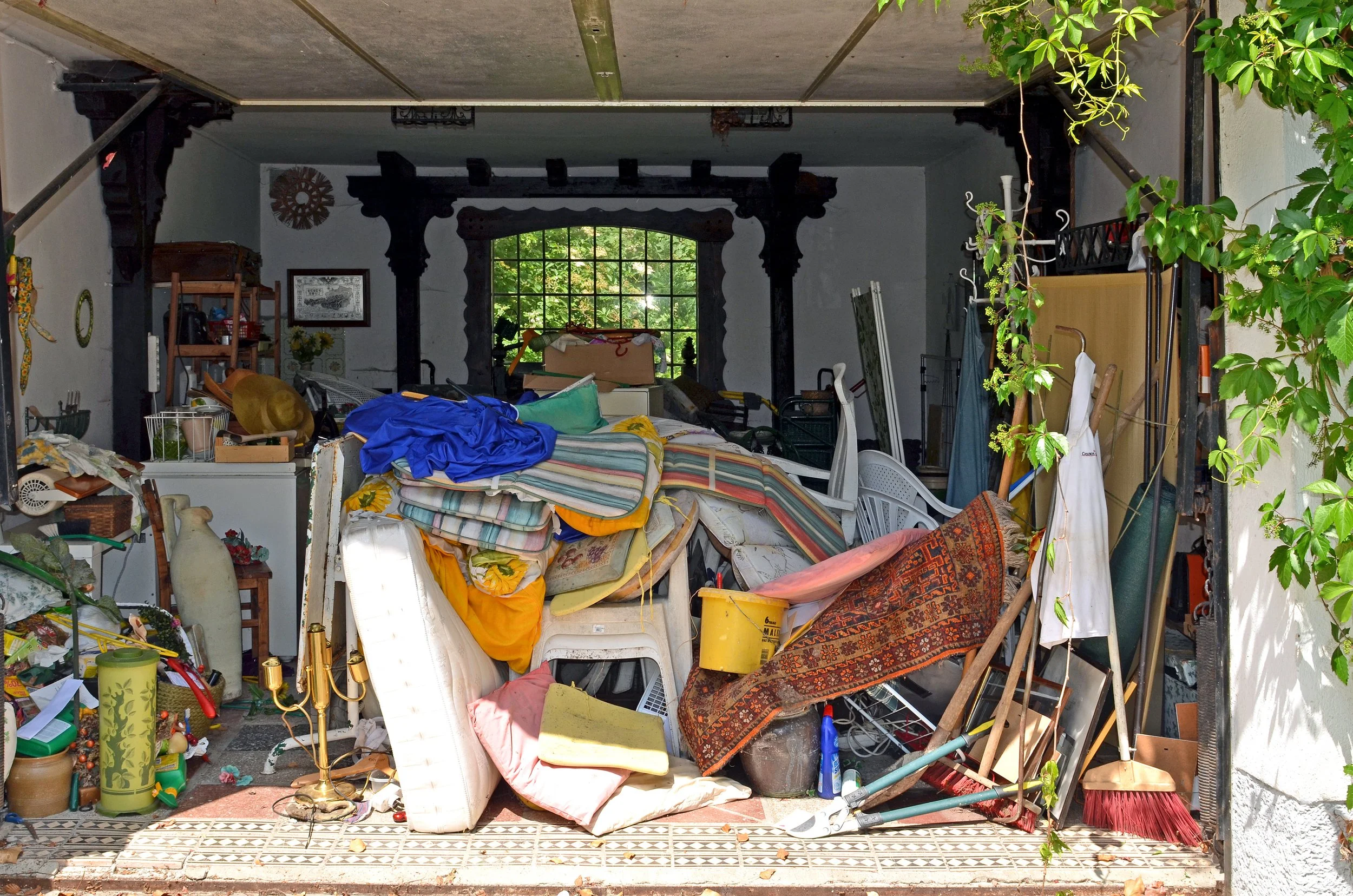 Messy garage filled with various clutter including furniture, fabric, and household items, with greenery visible through the window in the background.