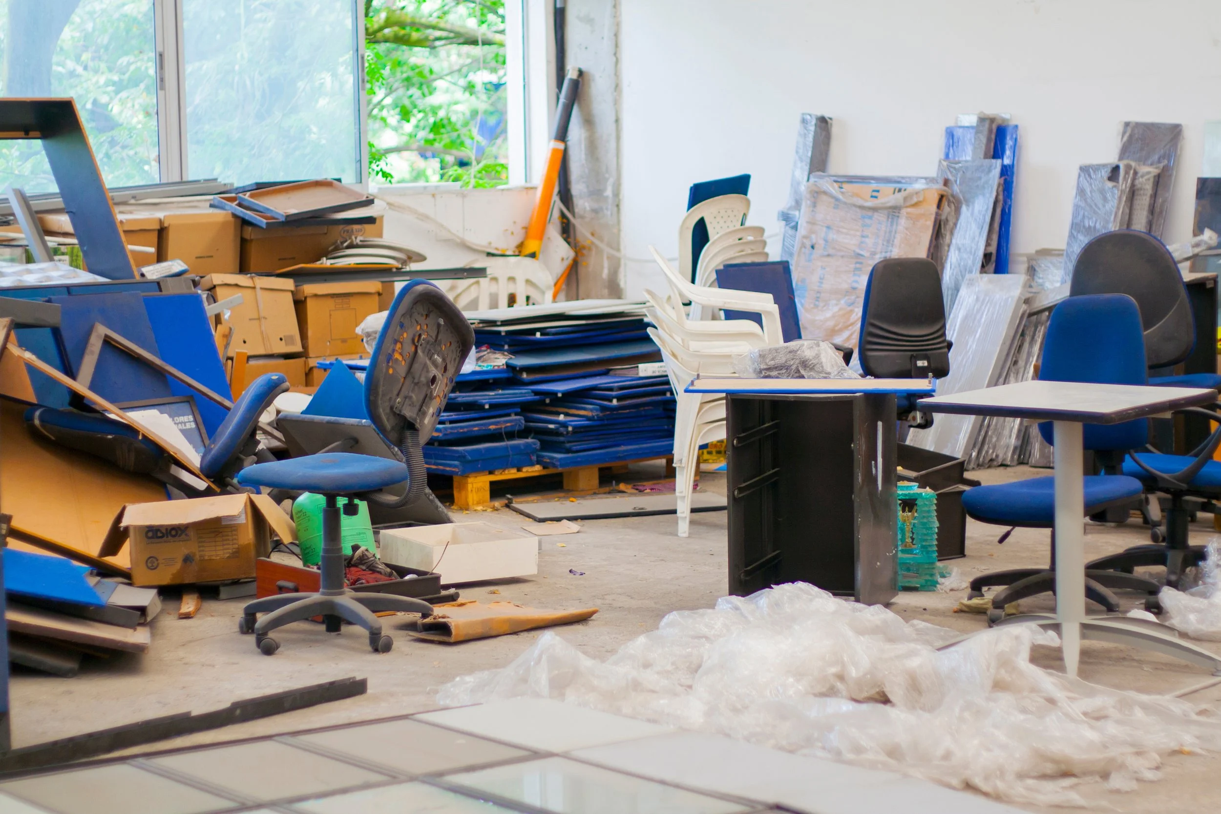 A cluttered office or workspace with chairs, desks, boxes, and various office supplies scattered around. There are broken or disorganized items, and the room appears to be in a state of disarray.