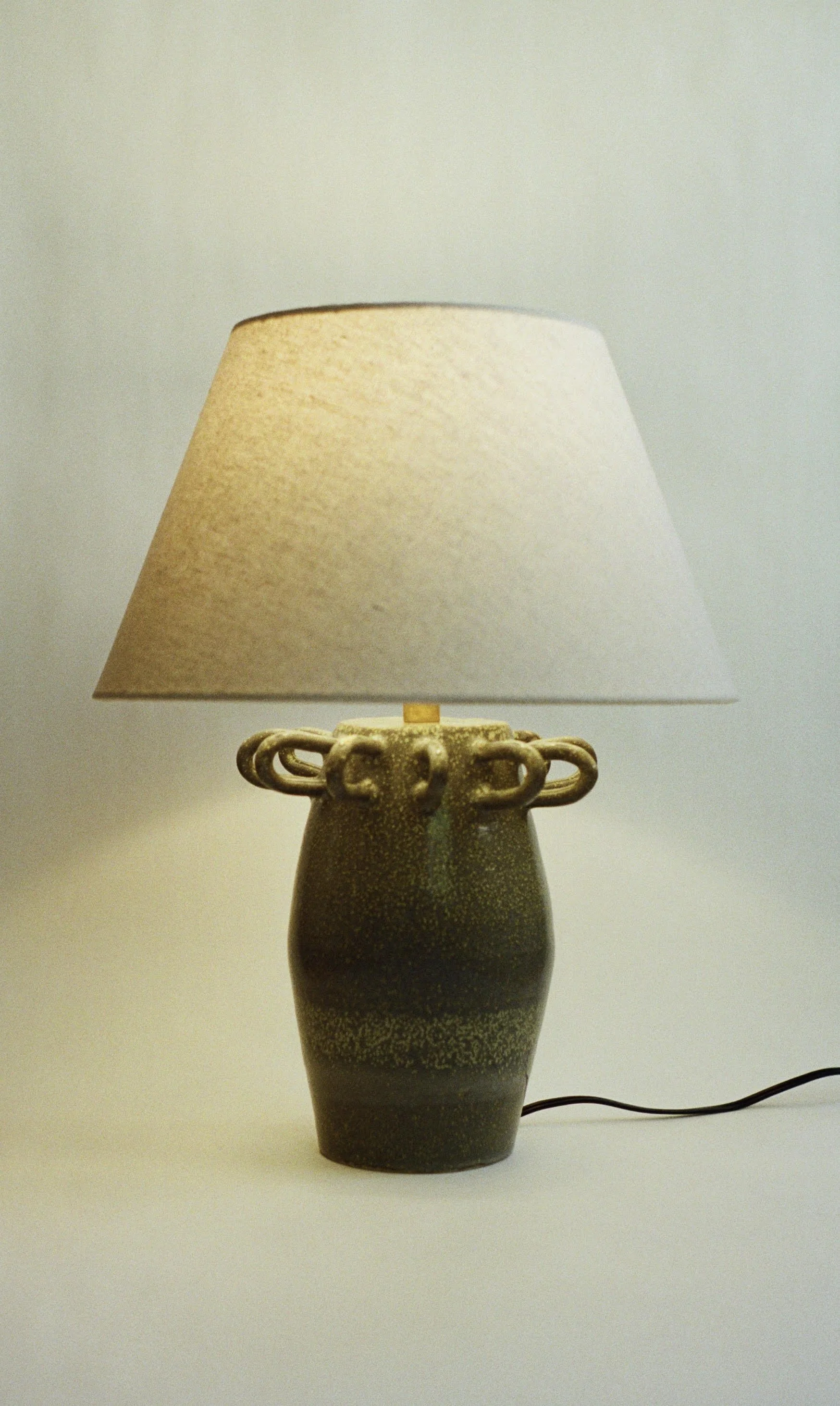 A ceramic table lamp with a textured beige lampshade and a dark speckled base, with a black electrical cord visible on the right side.