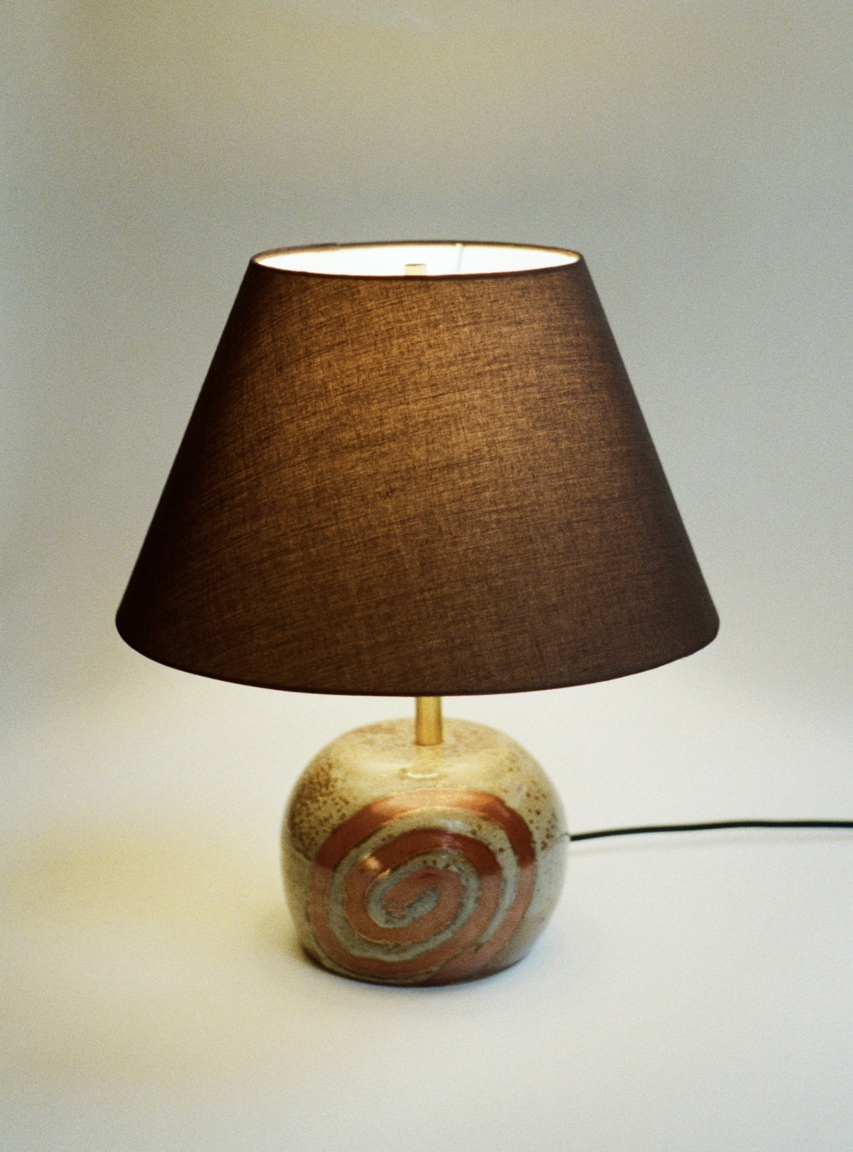 A table lamp with a beige base featuring concentric circles painted in red and gray, and a dark brown lampshade, placed on a white surface with a plain light-colored background.