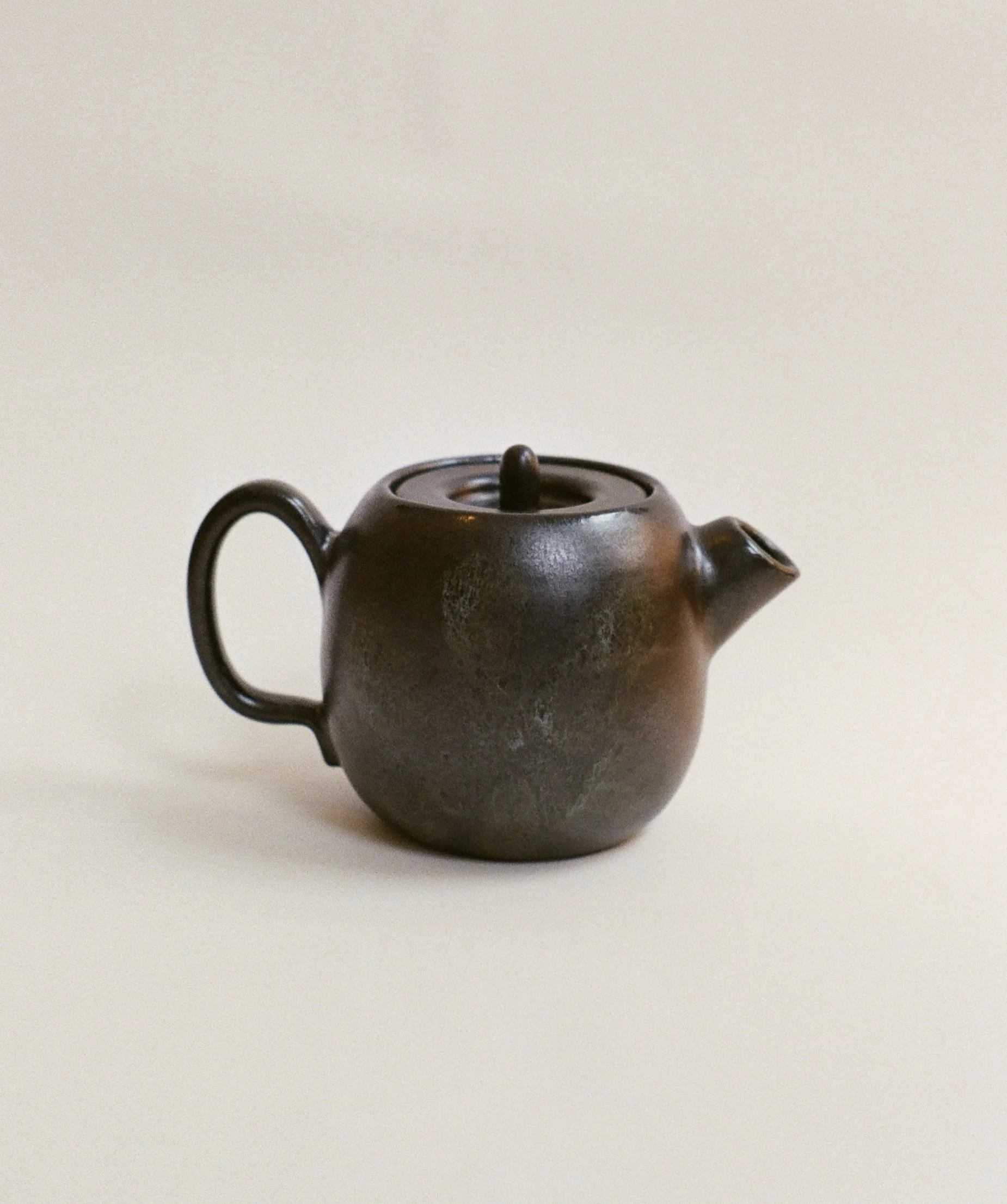 A small, round, brown ceramic teapot with a curved handle, a spout, and a lid, placed on a plain surface against a neutral background.