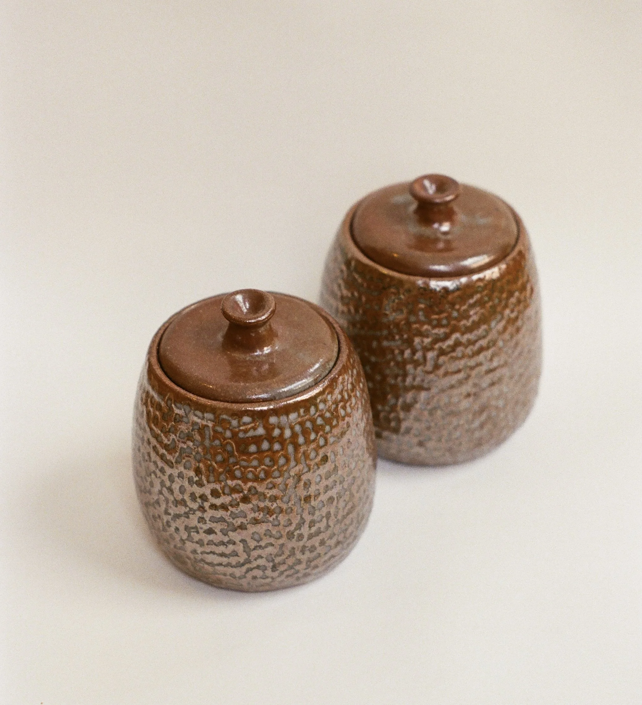 Close-up of three small, textured, brown ceramic containers with lids, placed on a white surface.