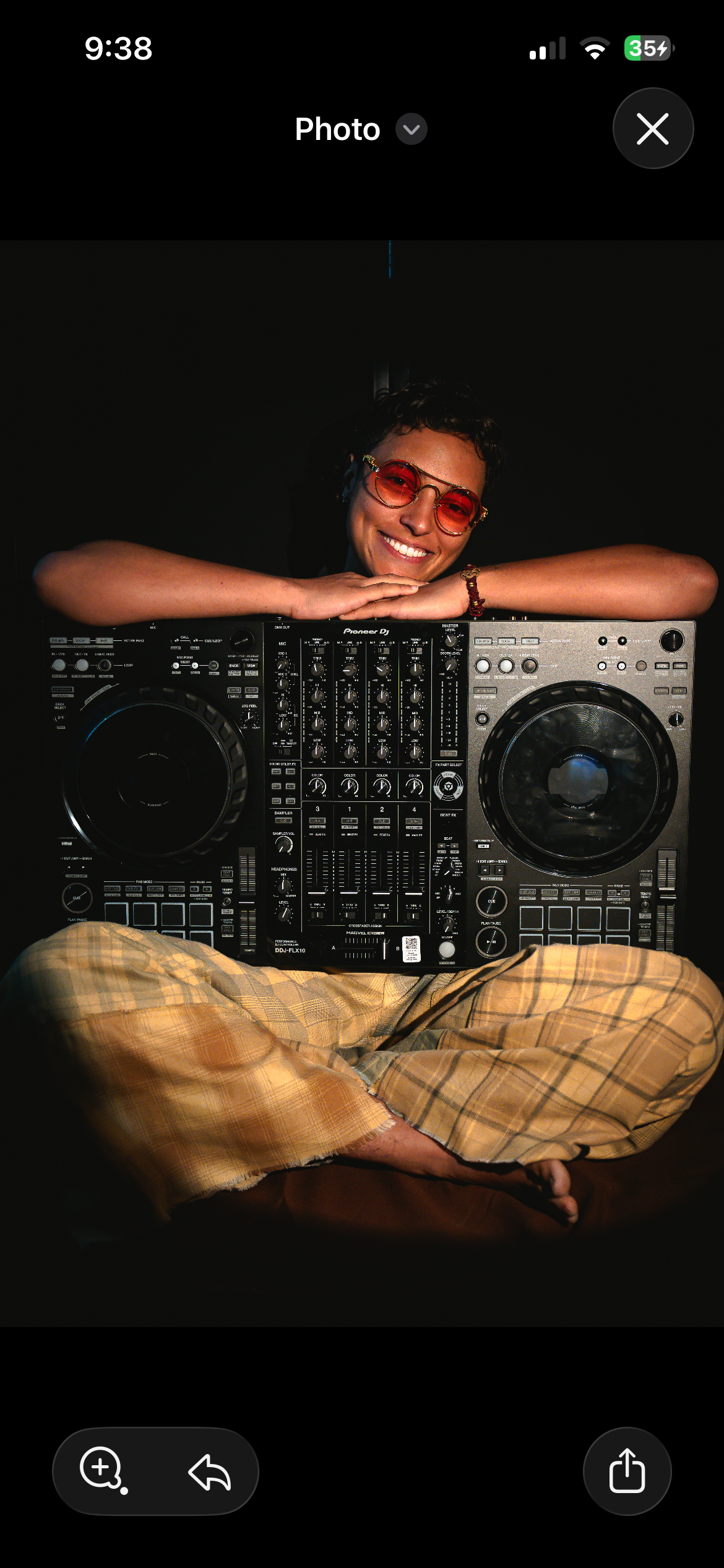 Person sitting cross-legged on the floor, wearing plaid shorts and sunglasses, smiling with arms resting on a DJ equipment setup including a Pioneer DJ controller.