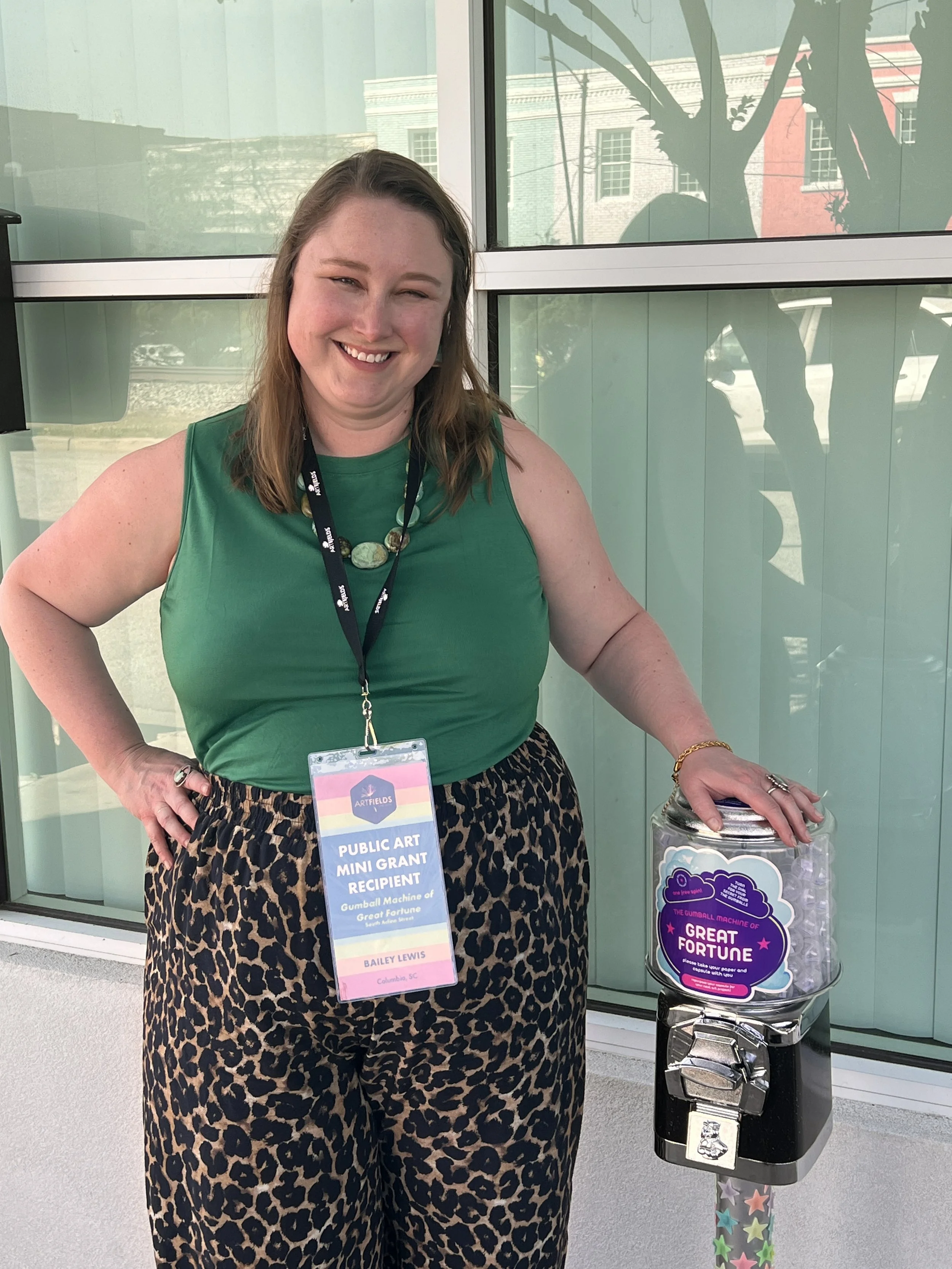 Bailey Lewis standing next to her Gumball Machine of Great Fortune public artwork, wearing an ArtFields artist badge identifying her and her public art as a 2026 ArtFields Public Art Grant recipient.