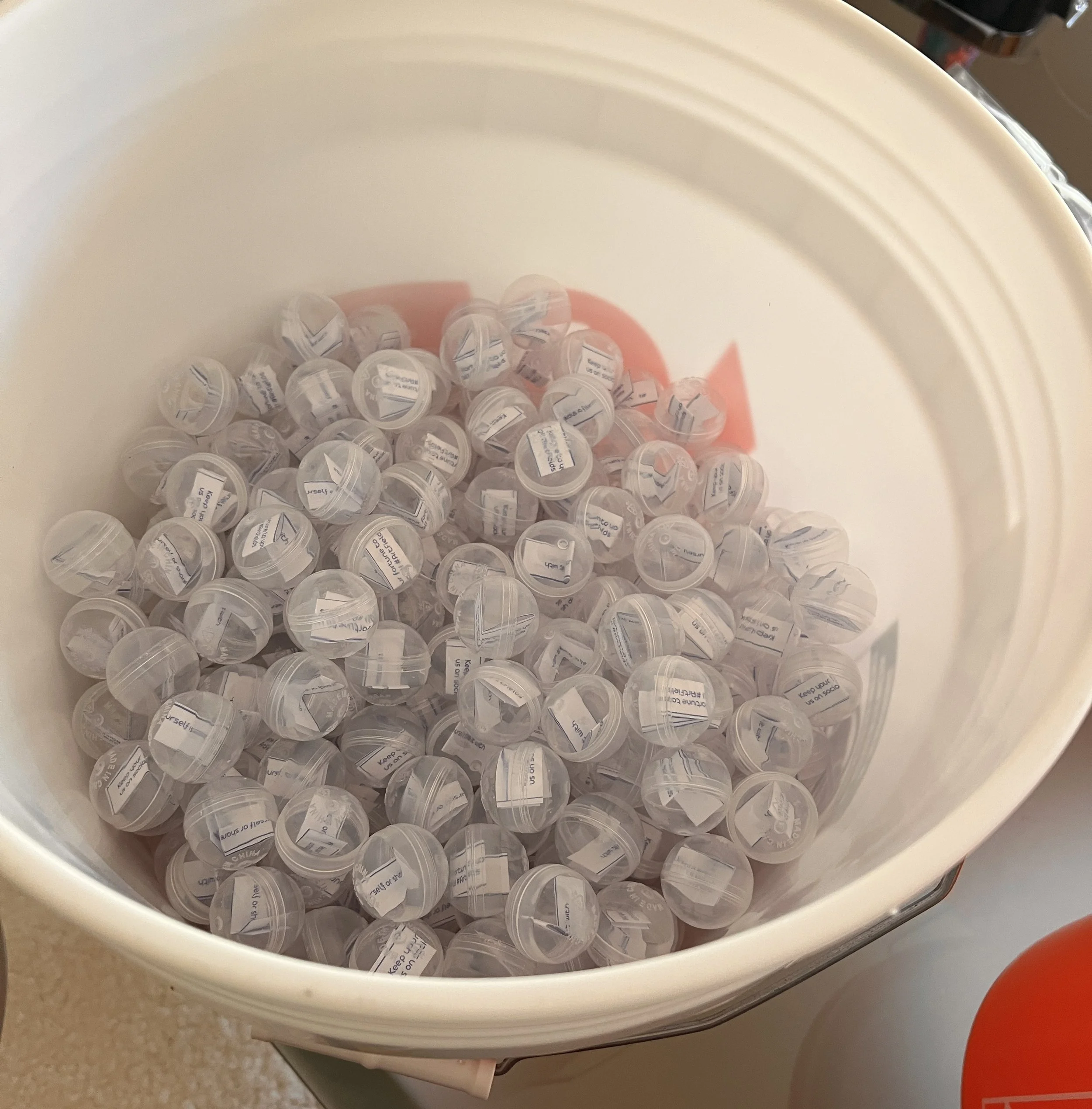Five-gallon bucket filled with small, clear capsules containing white paper fortune slips.