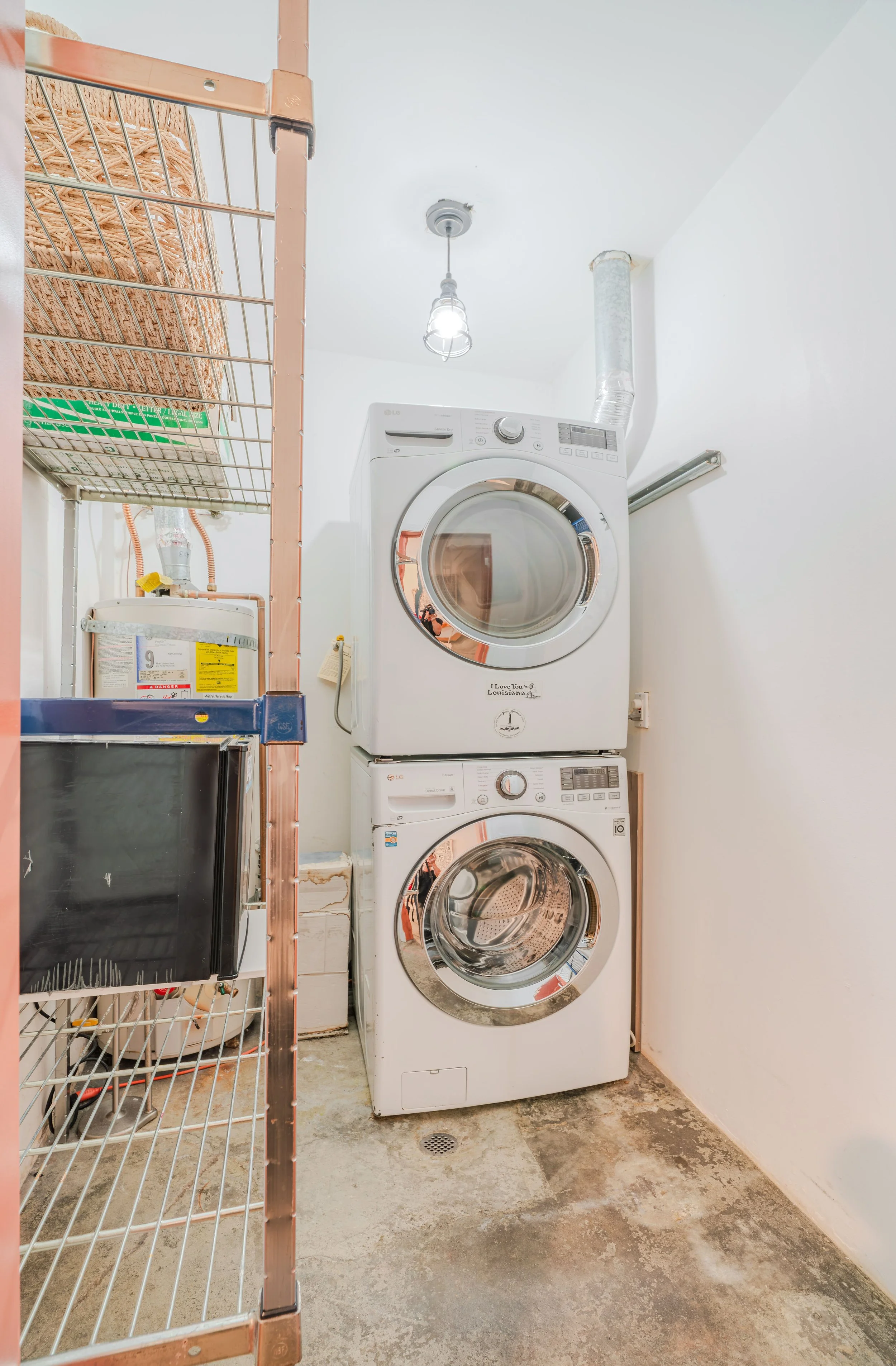 A laundry room with a stacked washer and dryer, a metal shelving unit with a wicker basket, a microwave, and a water heater against the wall. The floor is concrete and there is a drain on the floor.