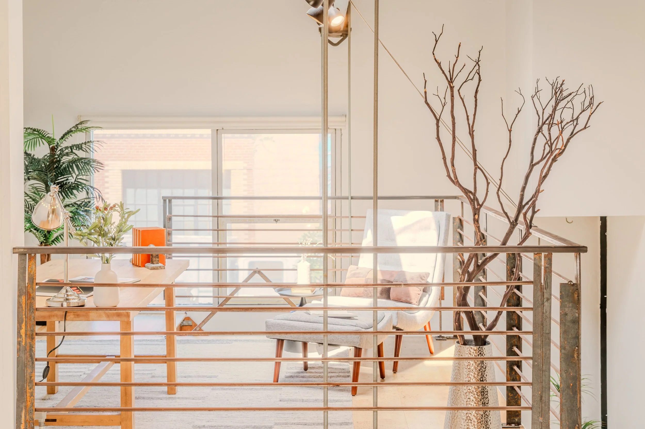 Interior view of a modern apartment or loft, showing a wooden dining table with a white ceramic vase and decorative objects, surrounded by gray upholstered chairs, with large windows bringing in natural light, a large potted plant, and decorative lea