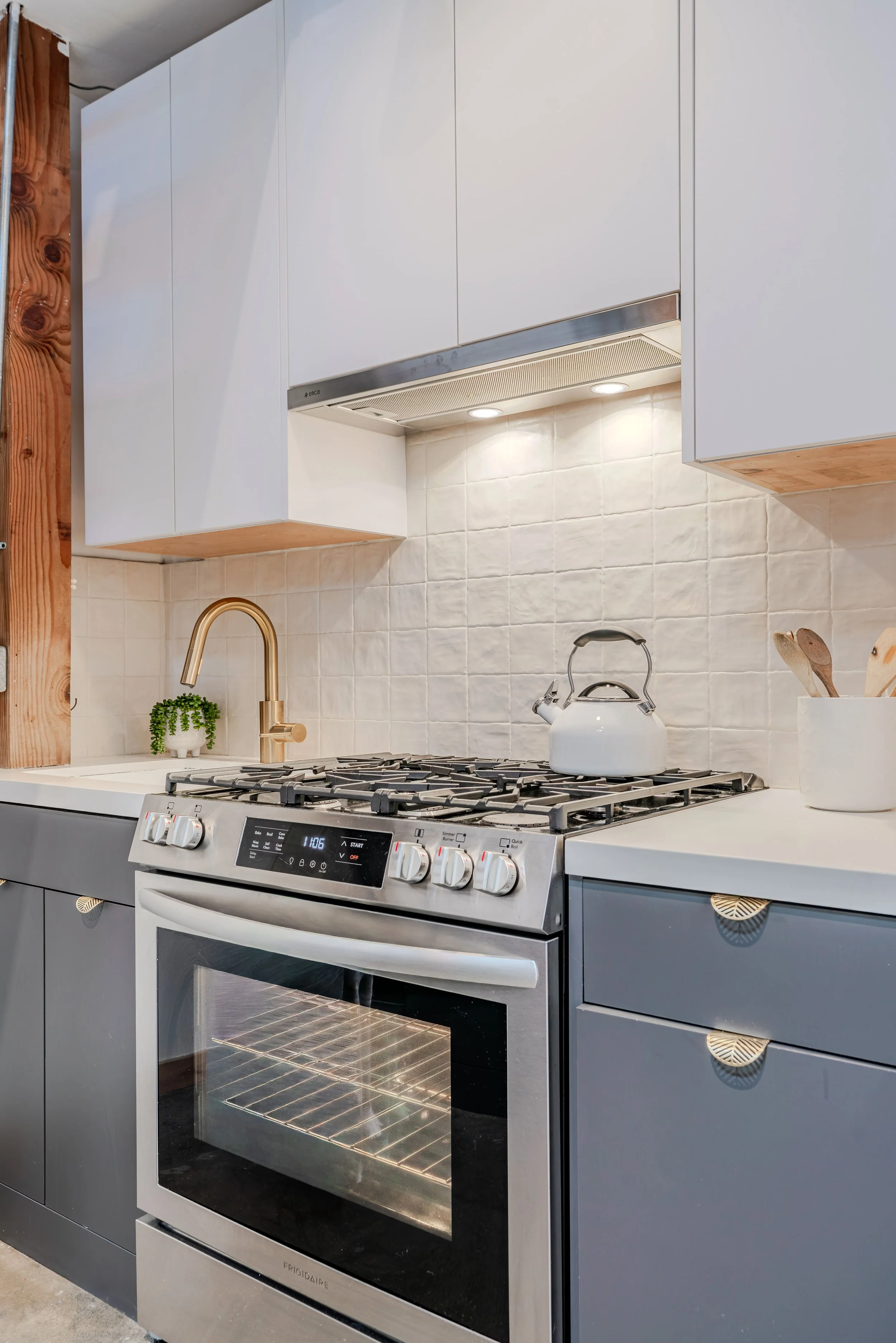 Modern kitchen with stainless steel oven, white upper cabinets, beige tiled backsplash, gold faucet, small green plant, white kettle, and wooden utensils on countertop.