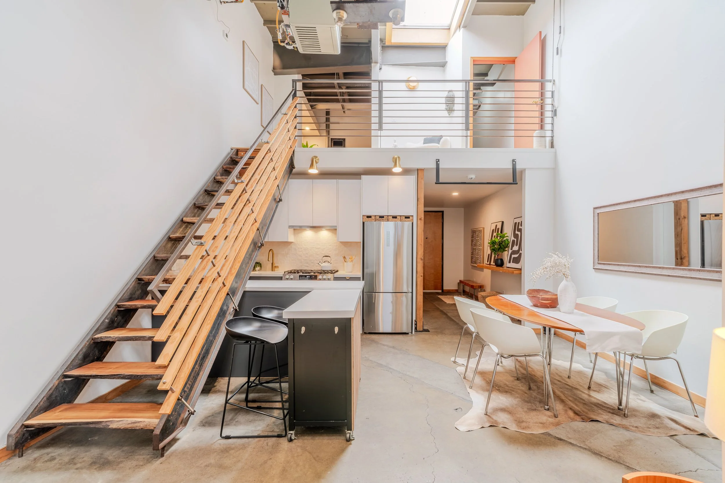 Open-concept modern loft with kitchen, dining, and living area, featuring a staircase with wooden steps and metal railing, white walls, and natural lighting.