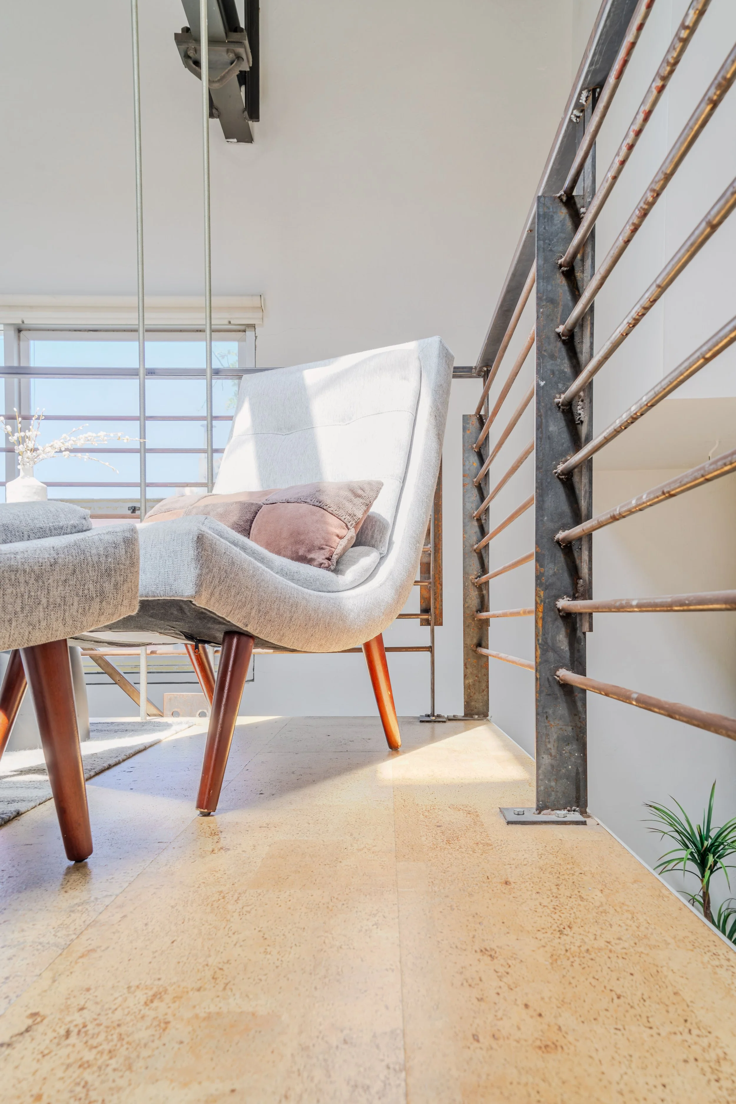 Living room with beige floor, white sofa with colorful pillows, metal railing, and a plant in the corner, lit by sunlight through large window.