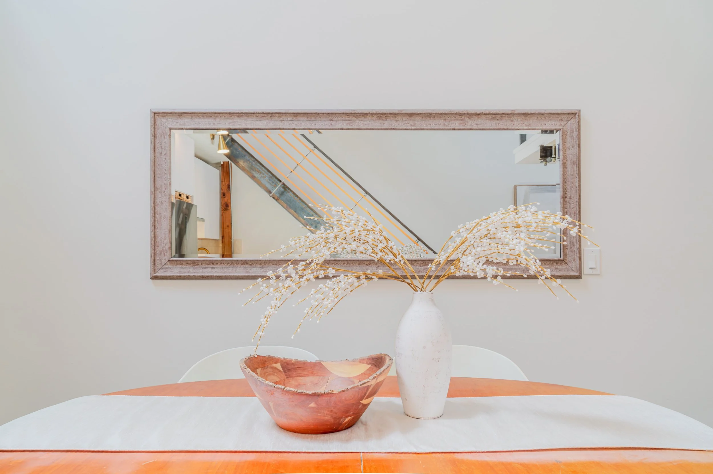A room with a large rectangular mirror on a white wall. In front, a table with a decorative vase holding dried branches and a wooden bowl.