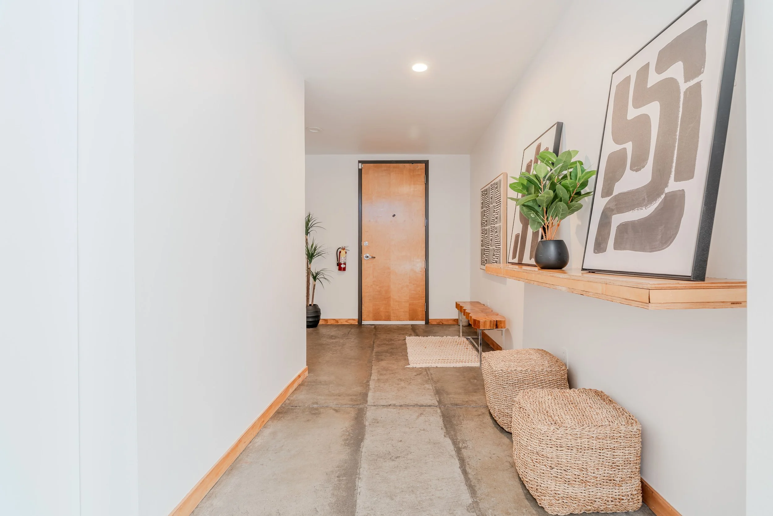 Interior view of a hallway with a wooden door at the end, art prints on the wall, potted plants, woven baskets, and a wooden bench.