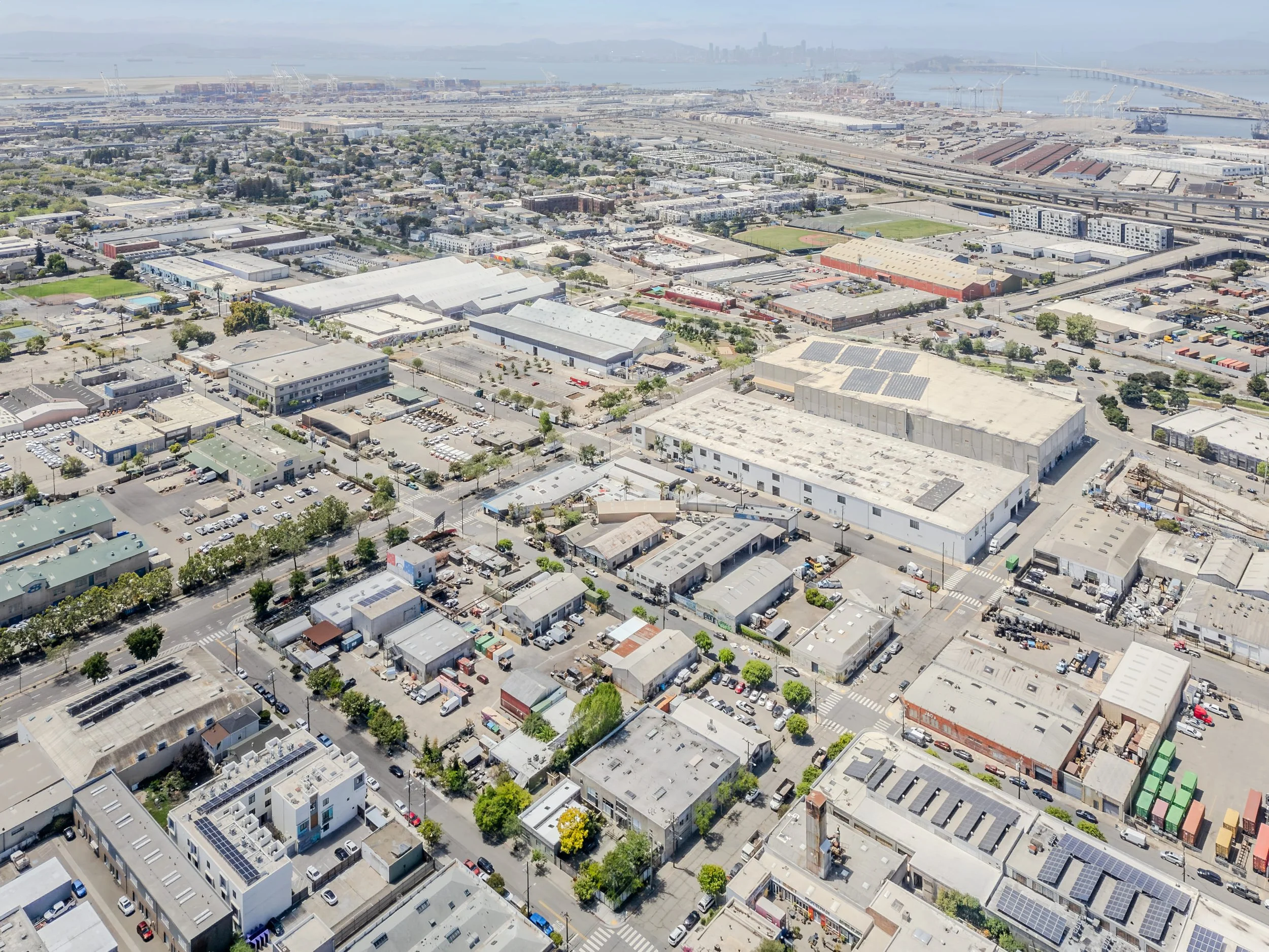 Aerial view of an urban area with various commercial and residential buildings, streets, parking lots, green trees, and sports fields, with a body of water and industrial port in the background.