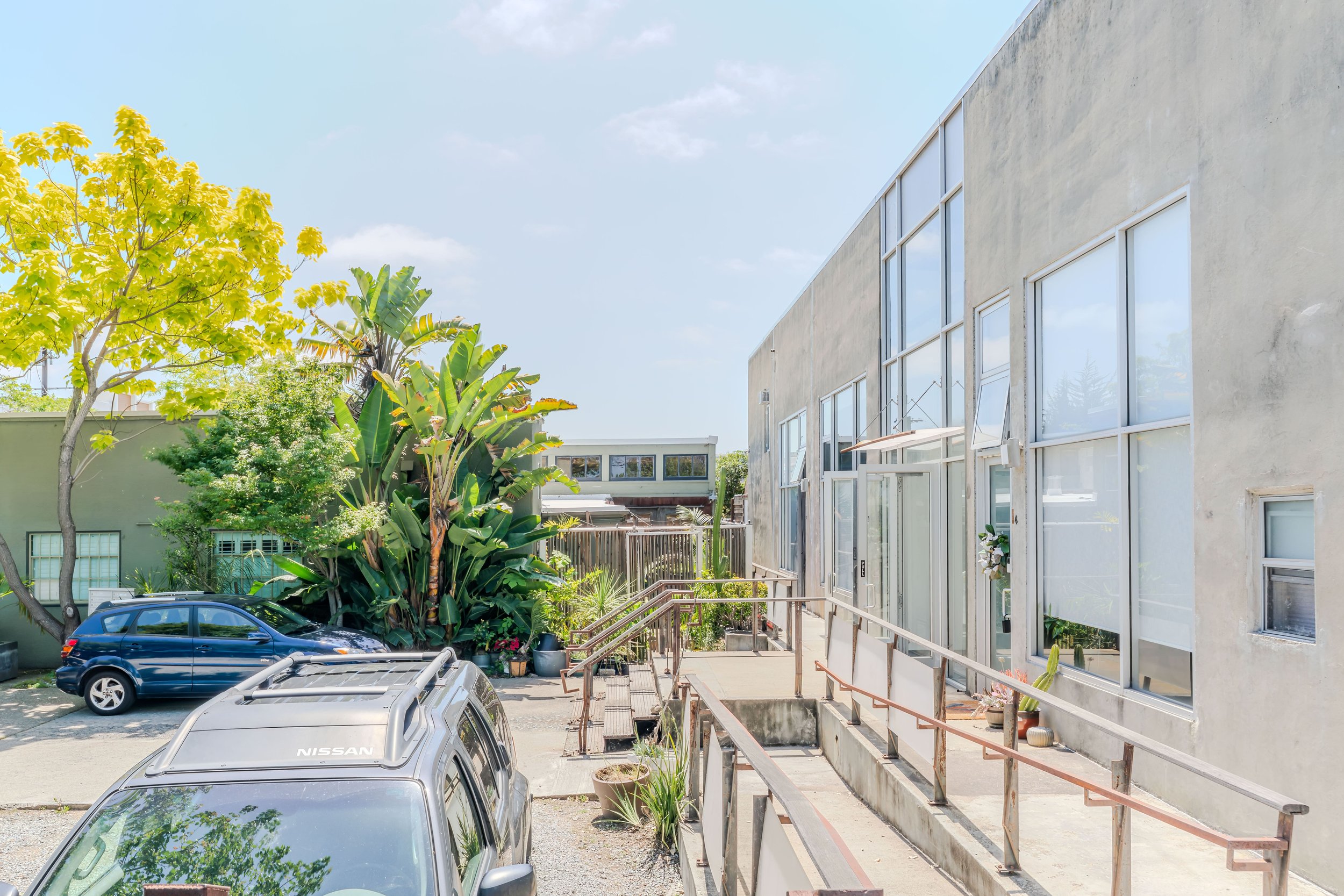 Parking lot with several cars parked, adjacent to a modern beige building with large windows, and a garden with trees and plants. Clear sky above.