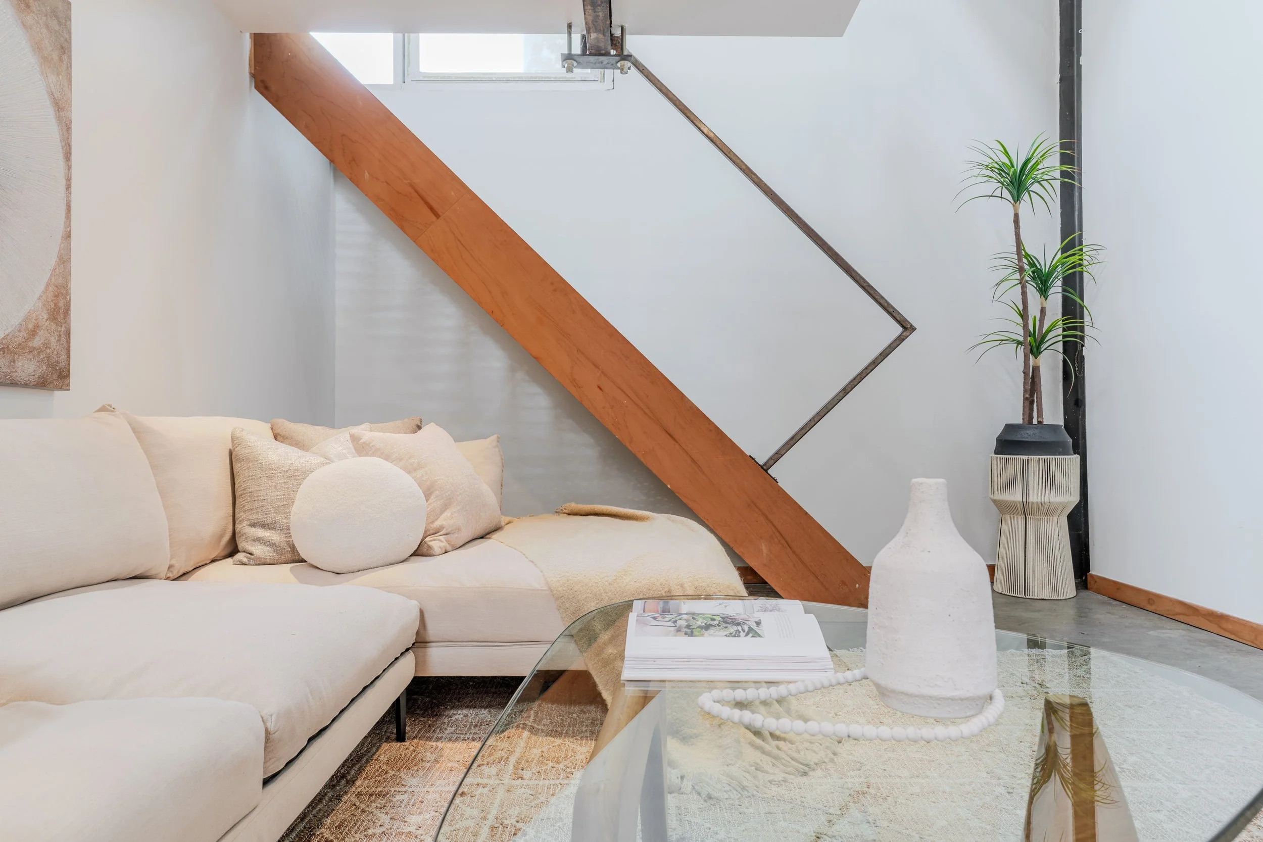 Living room with beige sofa, white cushions, glass coffee table with magazines, decorative vases, tall potted plant, and a staircase with wooden and metal railing in the background.