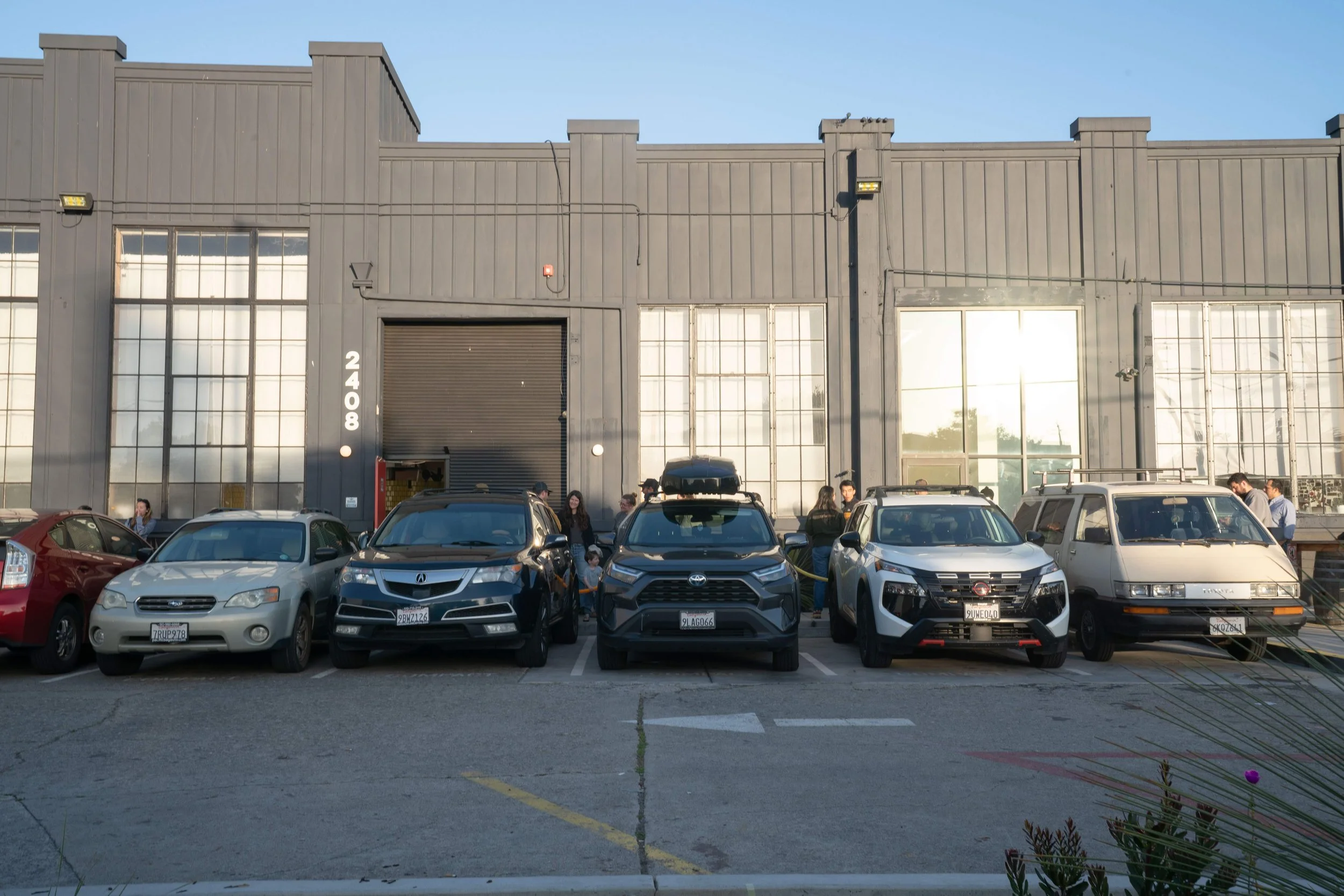 A parking lot in front of a large industrial building with large windows and a gray facade. Several cars are parked, and people are gathered near the entrance, some entering and exiting the building.