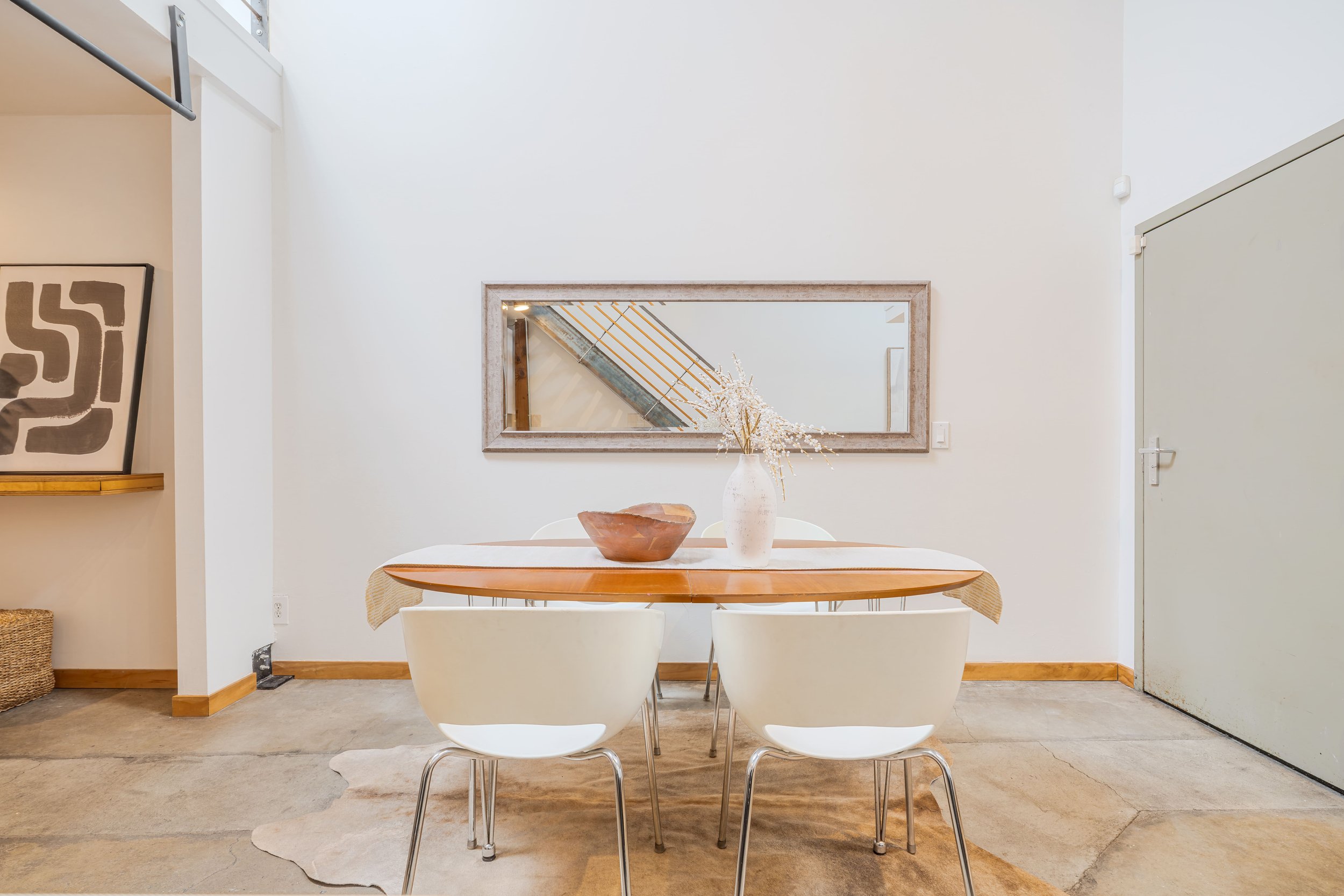 A dining area with a round wooden table, four white chairs, a decorative vase with dried flowers, a brown bowl, a large rectangular mirror on the wall, and a staircase reflected in the mirror, with white walls and light wood flooring.