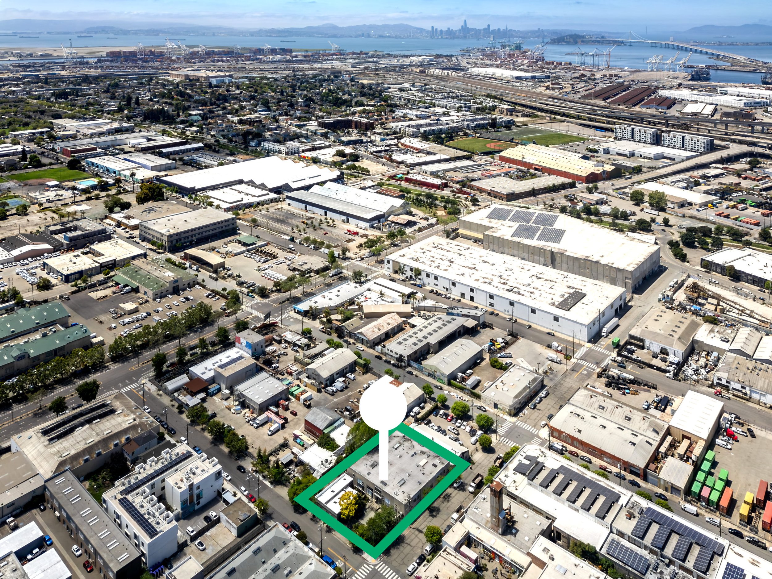 Aerial view of an urban area near a bay with cranes, ships, and a bridge in the background, showcasing industrial and residential zones, with a sports field and various buildings in the foreground.
