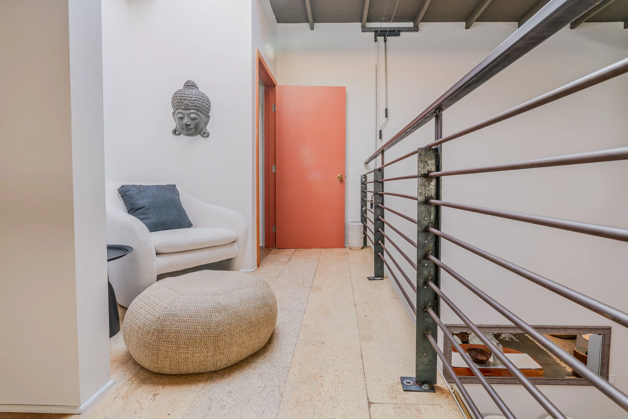 Interior of a modern home with a white sofa, gray pillow, round woven ottoman, a coral-colored door, a ceramic face sculpture on the wall, and a metal railing along a staircase.
