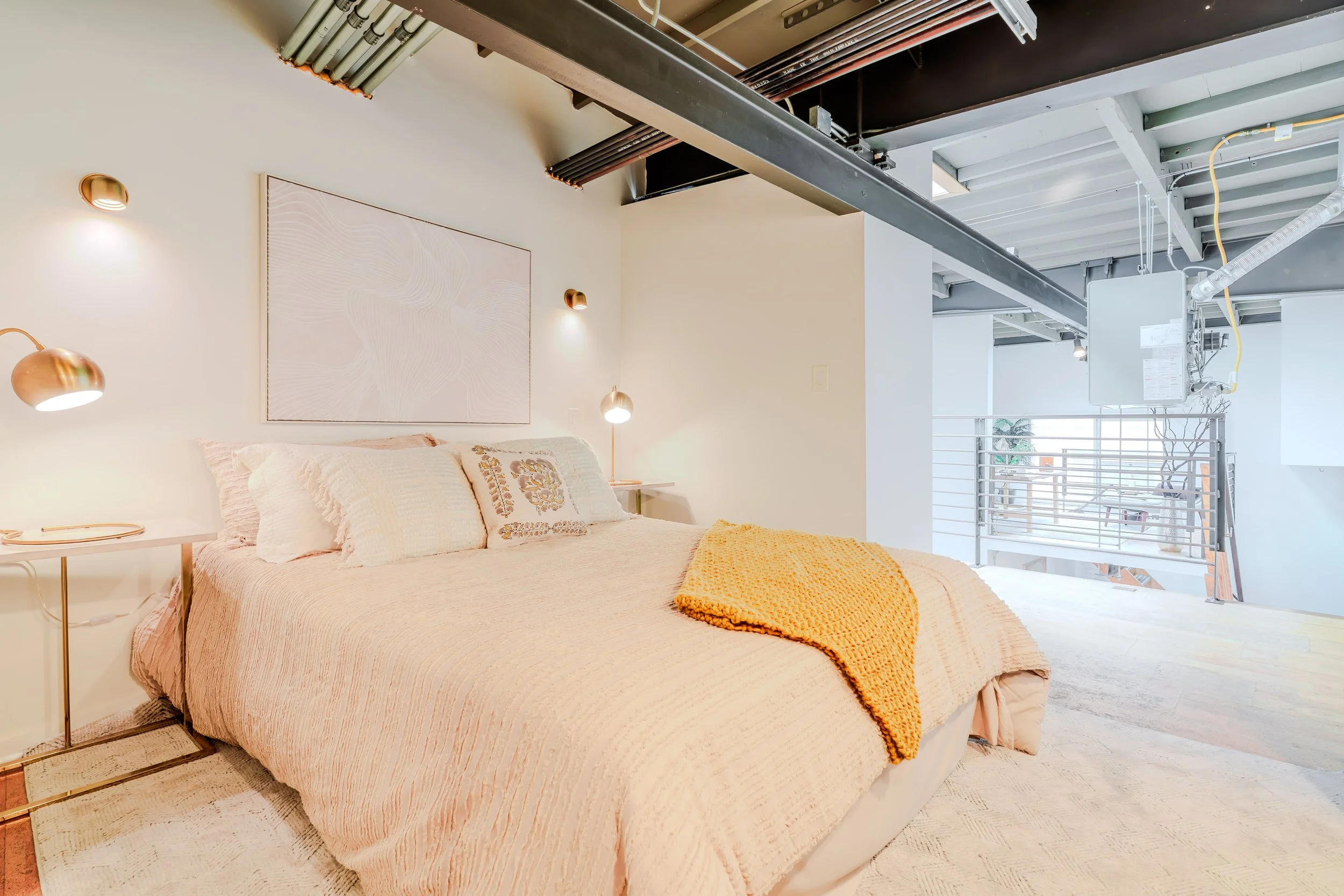 A modern bedroom with a bed covered in a light-colored blanket and pillows, a side table with a gold lamp, and a large wall art piece above the bed. The room has white walls and ceiling, with exposed industrial-style ceiling beams and ductwork, indic