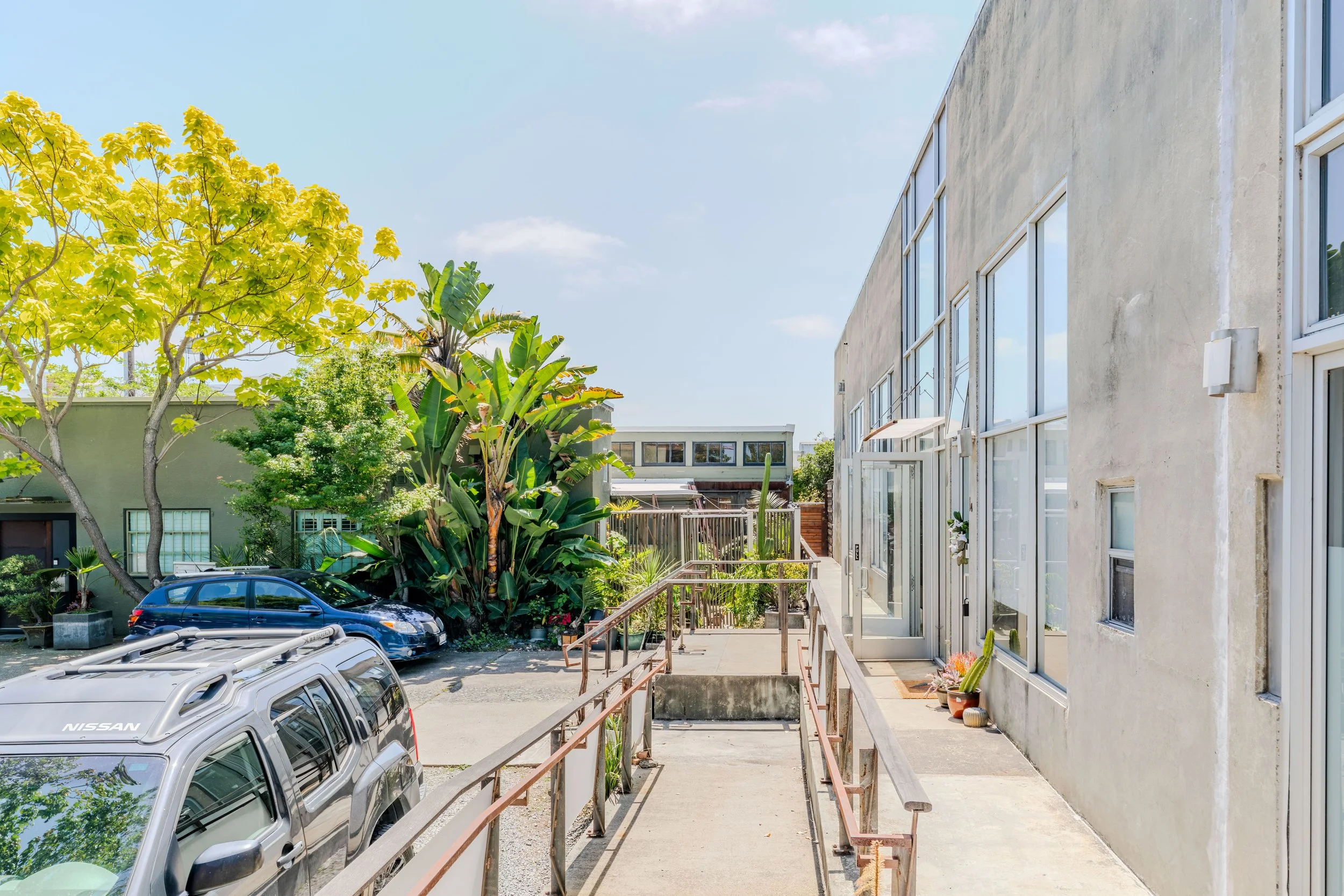 Apartment building with a sidewalk and a small parking lot filled with several cars. There are trees and plants along the side of the building, and the sky is clear with a few clouds.