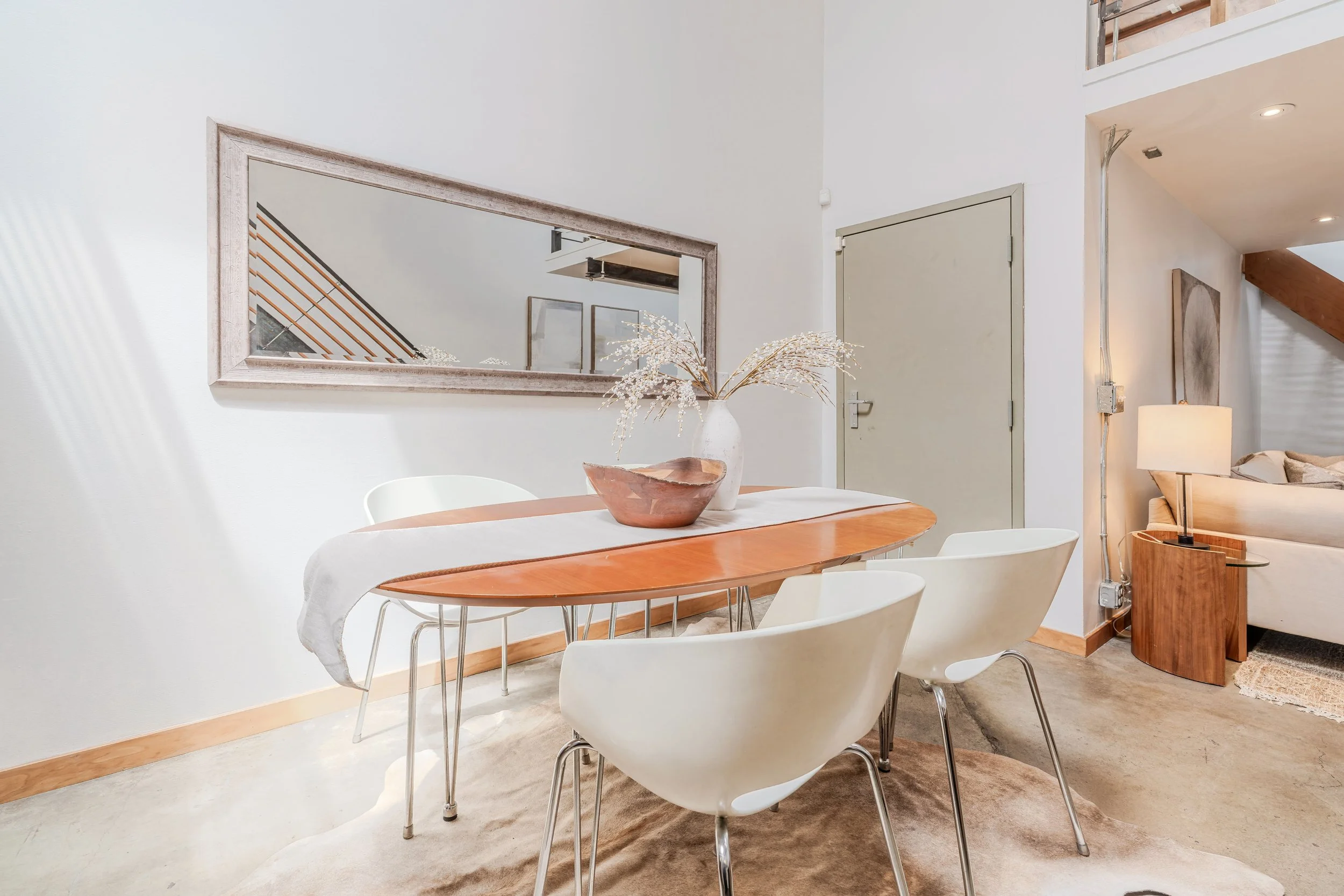 The image shows a dining area with a round wooden table covered by a white tablecloth, surrounded by white modern chairs. A vase with white dried flowers and a wooden bowl are on the table. A large mirror with a wooden frame is mounted on the wall ab