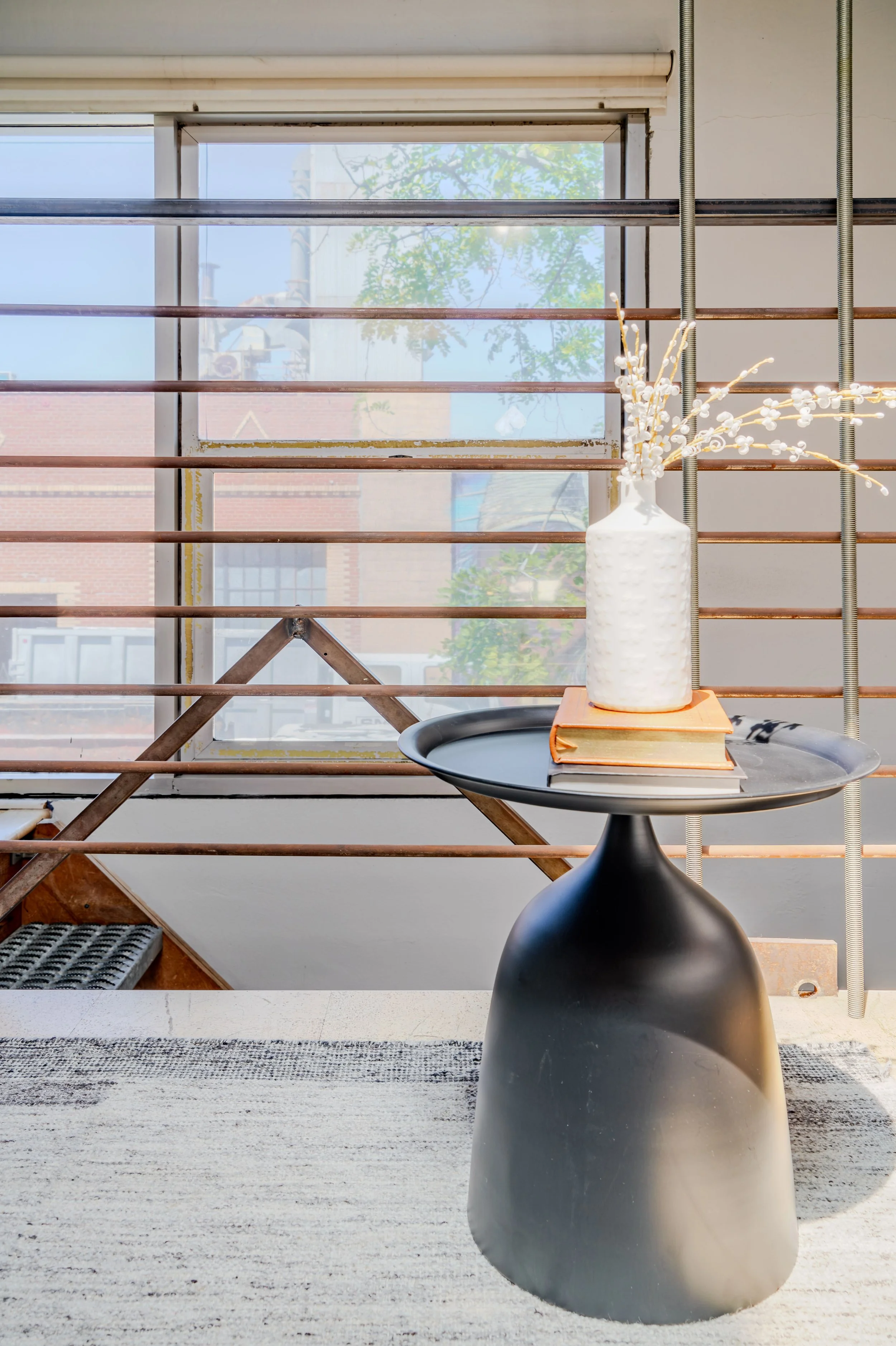 A black vase with textured surface on a round black table, holding white flower branches, in front of a window with horizontal blinds, in a room with a striped rug on the floor.