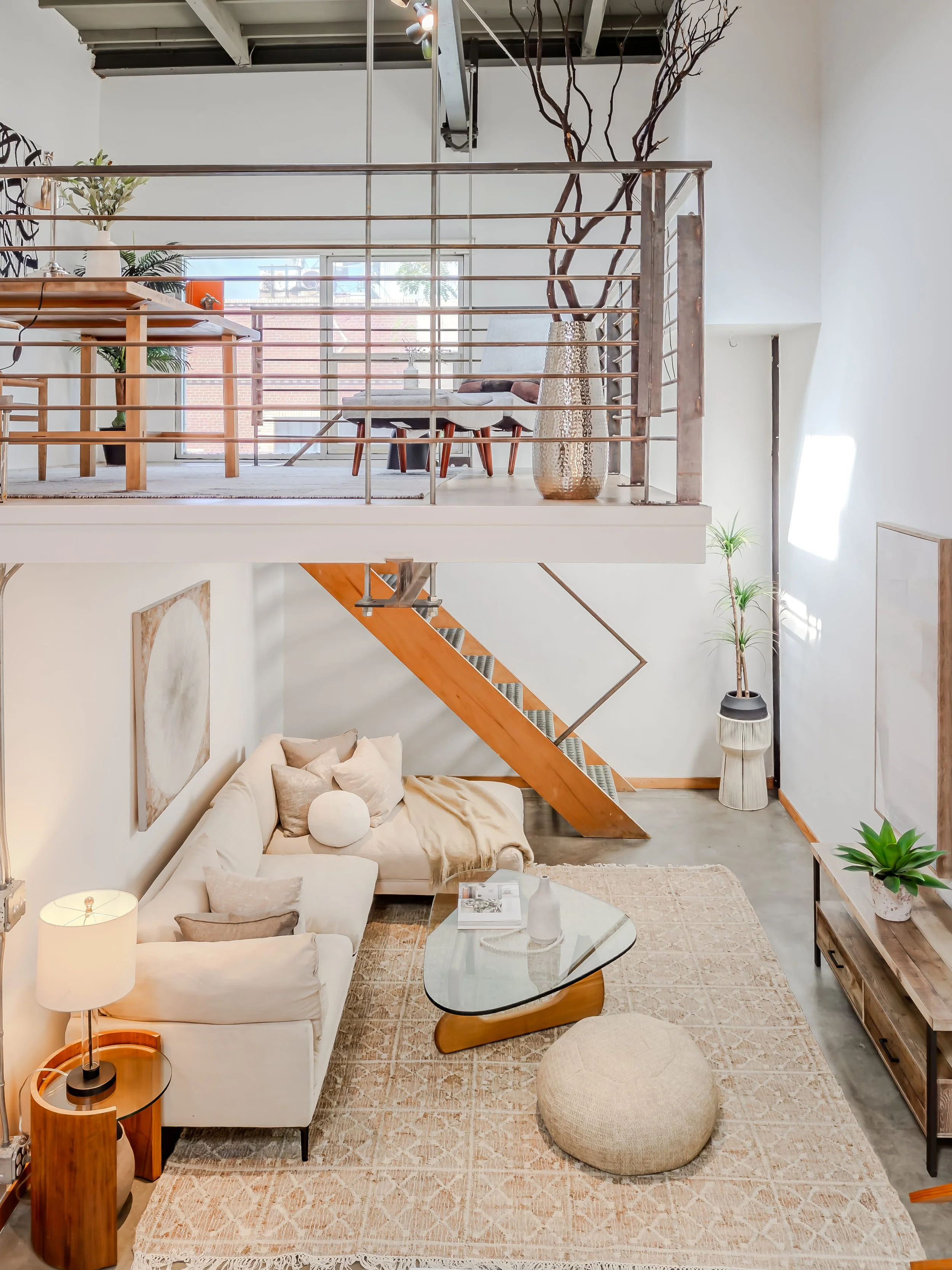A modern living room with a white sofa, a glass coffee table, a beige rug, and a wooden side table with a lamp. There is a large artwork on the wall, a potted plant, and a cozy pouf. An upstairs dining area with a table, chairs, and decorative plants