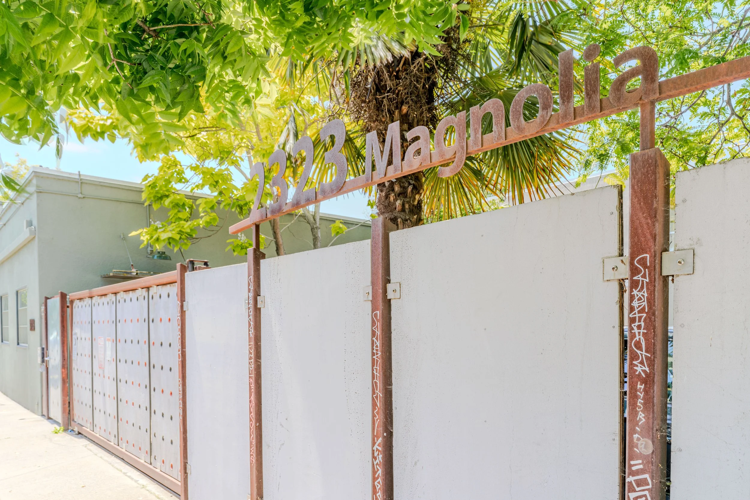 Street scene with a metal sign displaying the date '32 3 May' above a concrete wall and a property fence, surrounded by green trees and a building in the background.