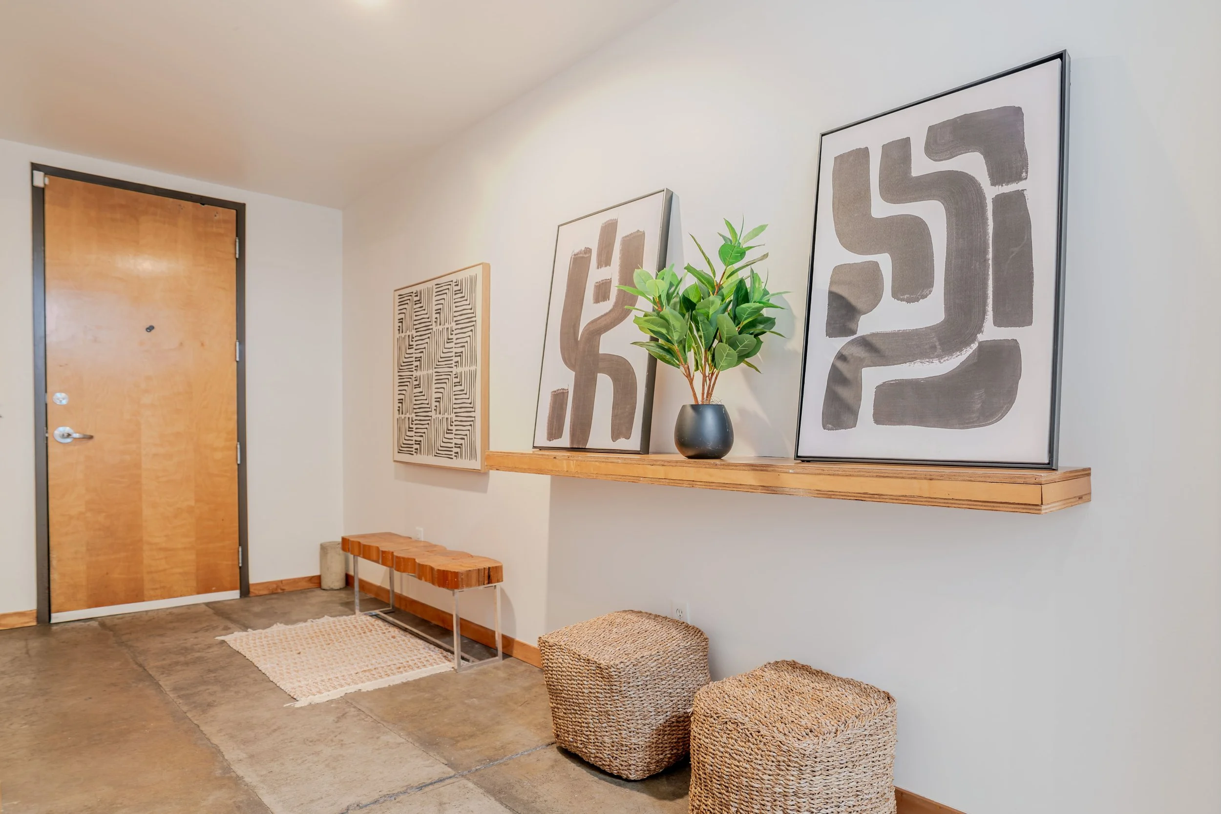 Decorative wall art with black and white abstract designs, a green potted plant on a wooden shelf, twin woven ottomans, a small woven rug, near a wooden door in a minimalist hallway.