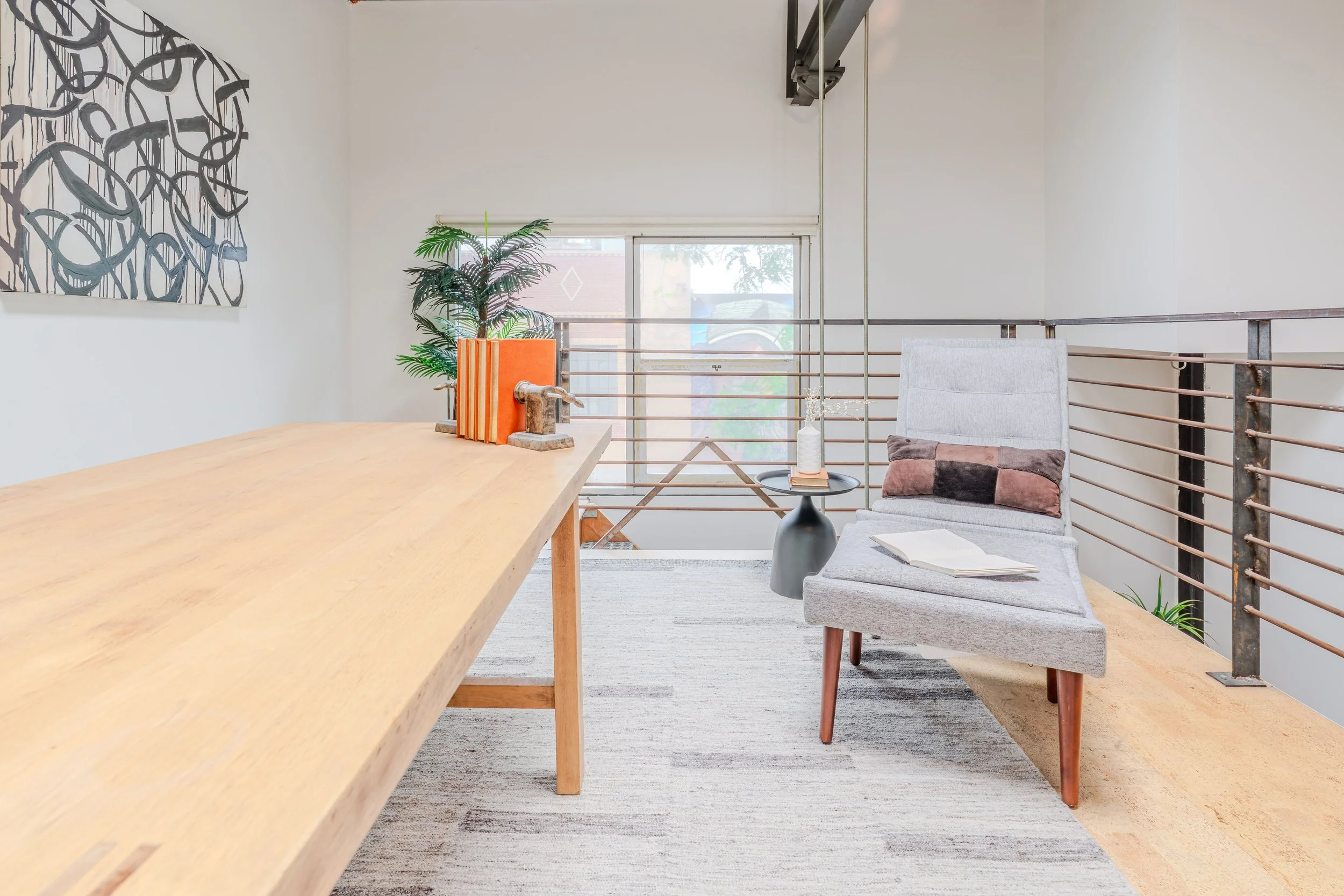 A bright indoor space with a large window, a modern armchair with a multicolored throw pillow, a small round table with a decorative vase, a potted plant, and minimal wall art.