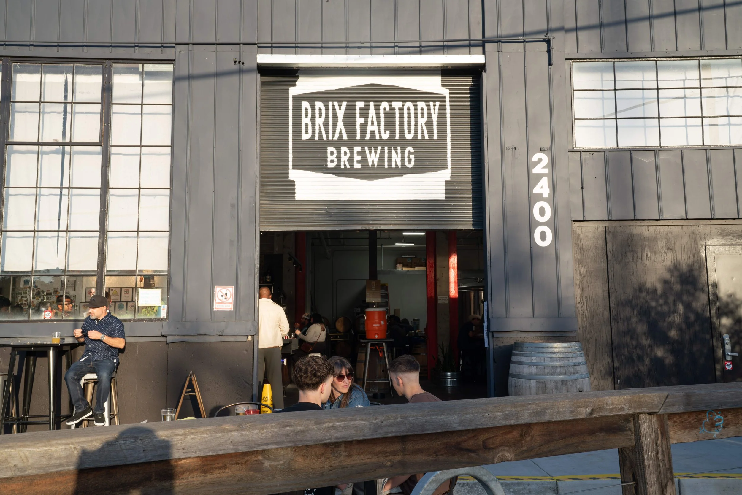 Exterior view of Brix Factory Brewing with people sitting outside and inside, some standing, near large windows and a rolling door with the brewery's sign, street number 2400, on the building.