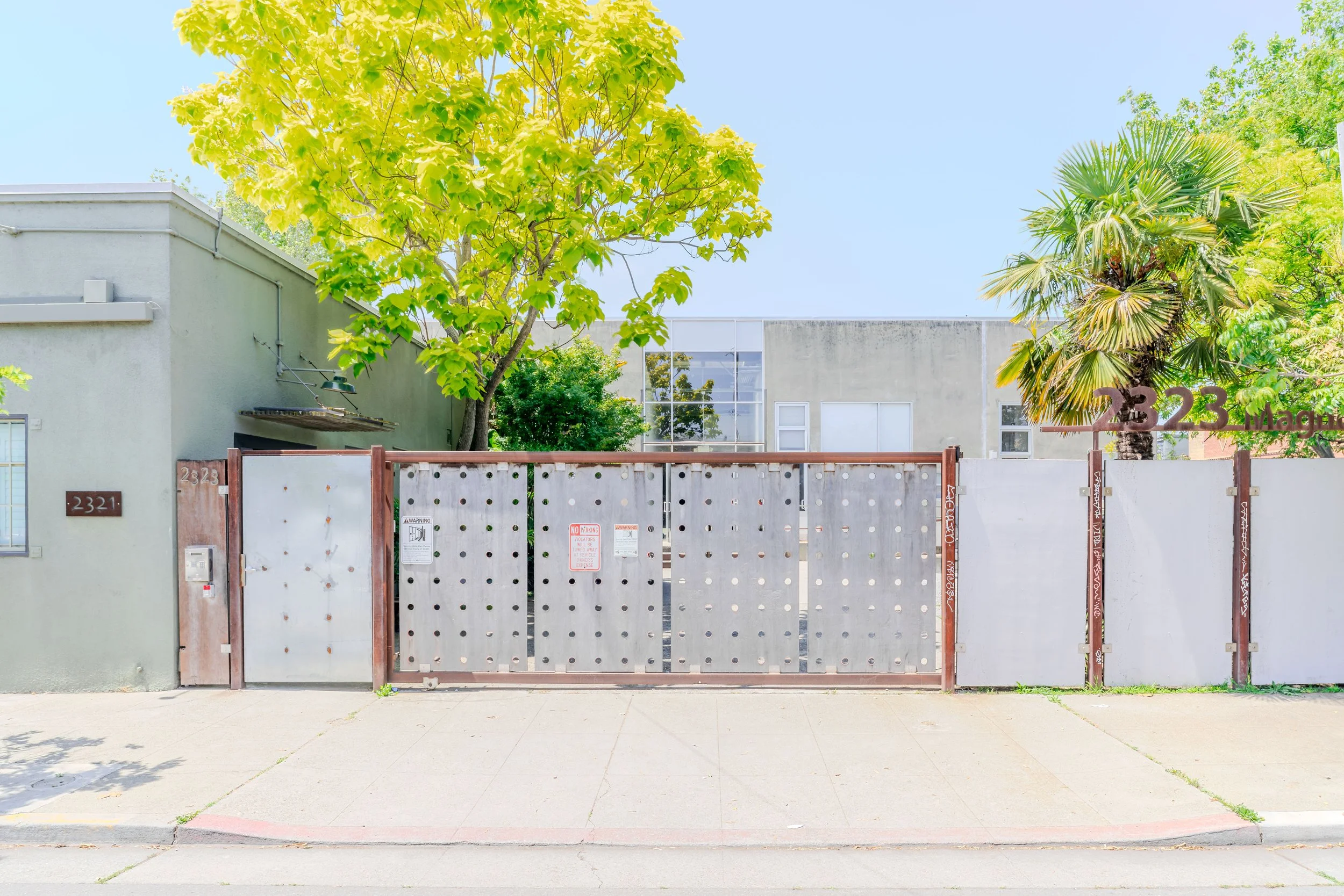 A closed metal gate in front of a modern building with a large tree and other greenery, under a clear blue sky.