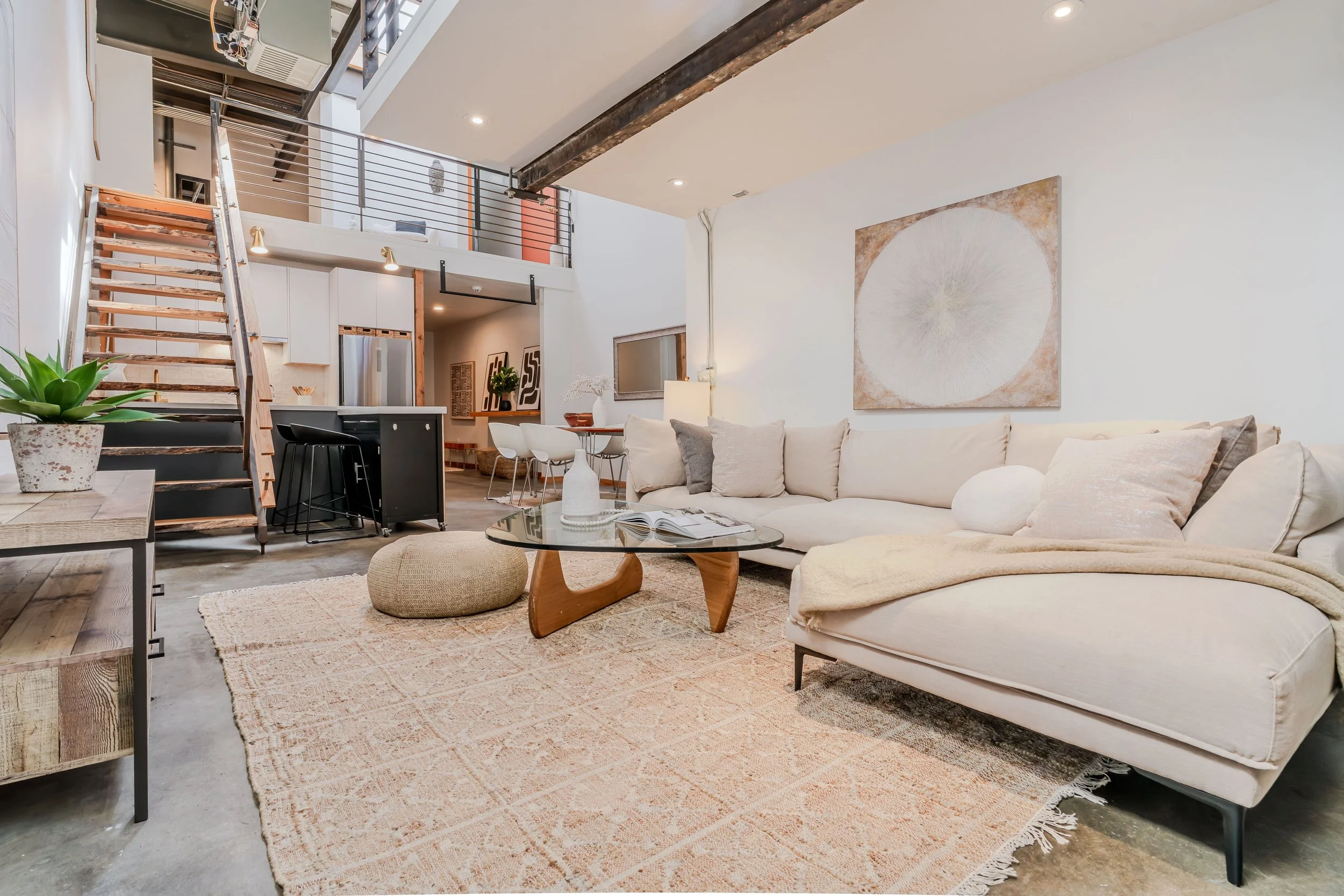 A living room with a cream-colored L-shaped sofa, a glass coffee table with a wooden base, and decorative pillows. There is a large abstract artwork on the wall and a rug underneath the furniture. The room has an open layout with a staircase leading to a loft area and an industrial-style ceiling with exposed beams.