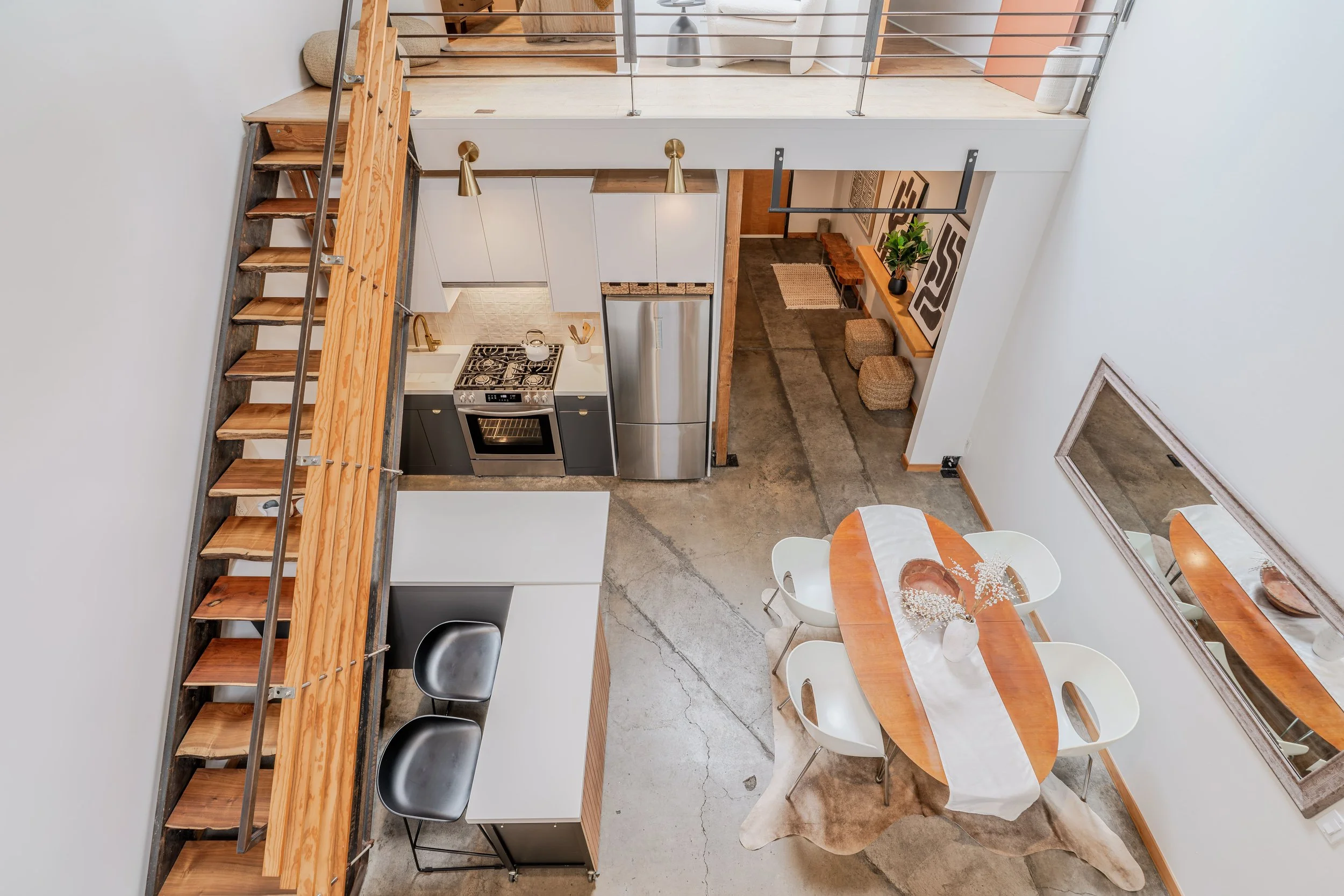 A view from above of a modern, open-concept kitchen and dining area with a staircase. The kitchen has white cabinets, a stainless steel refrigerator, gas stove, and black sink. The dining area features a wooden table with white chairs and a flower ar