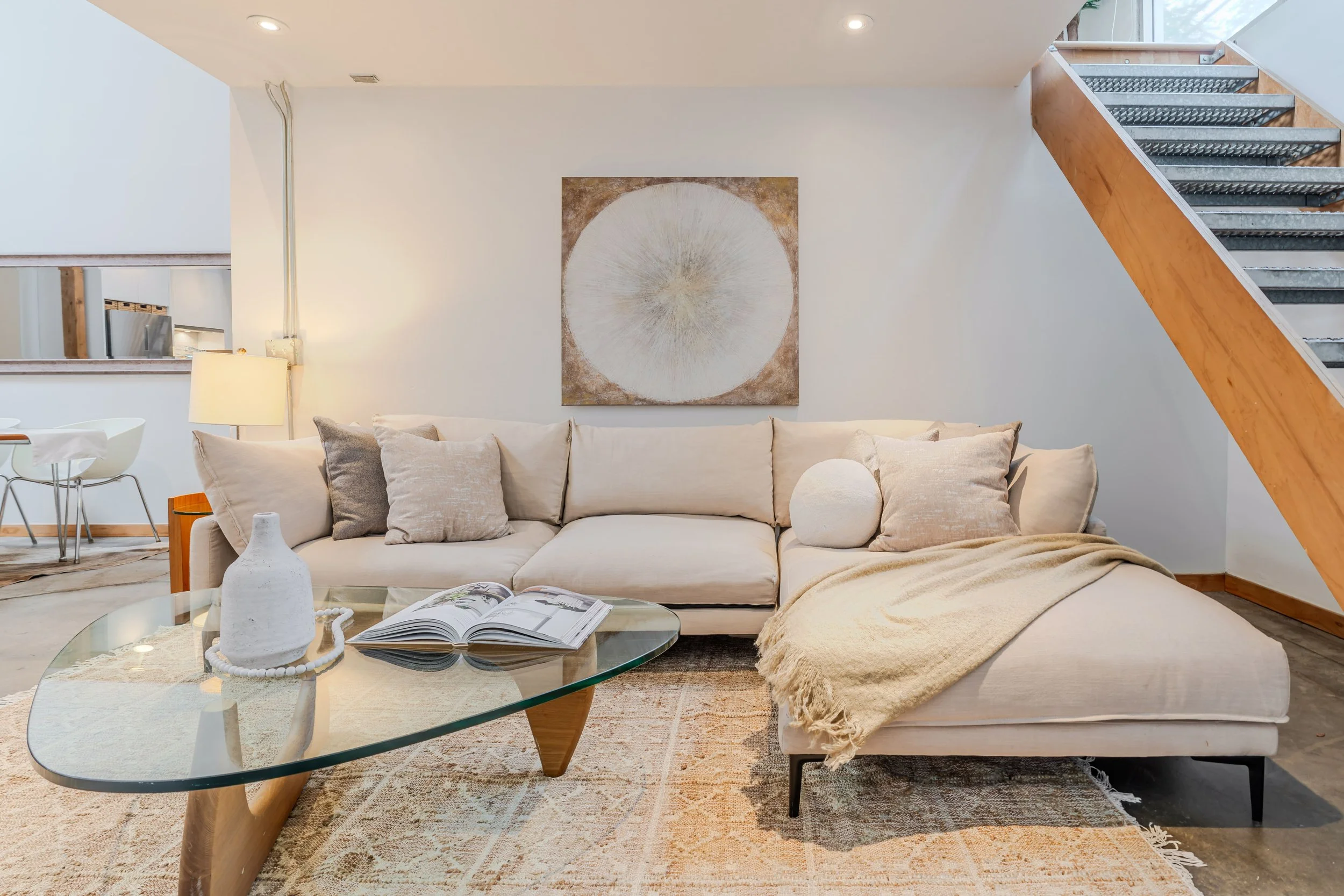 A modern living room with a beige sofa adorned with neutral pillows, a glass coffee table with decorative objects and an open magazine, a textured area rug, and a large abstract circle wall art above the sofa. To the right, a wooden staircase with me