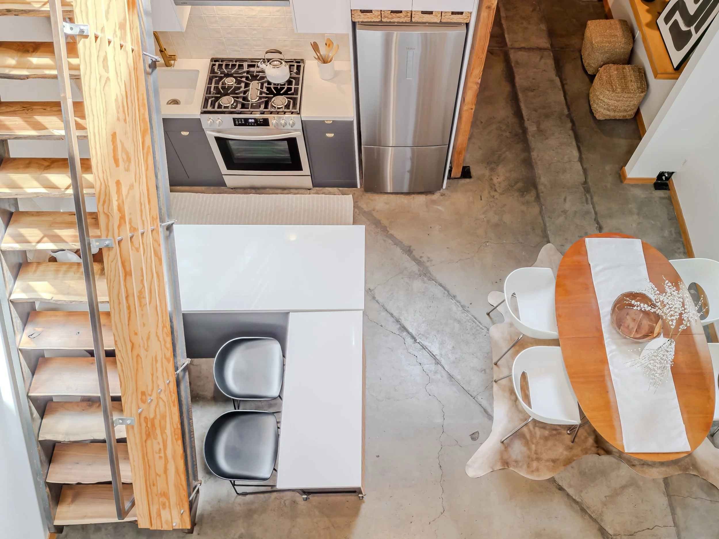 Top-down view of a modern kitchen with a stainless steel refrigerator, a gas stove with a kettle, white countertops, and a small dining table with four white chairs, set with a decorative centerpiece and cloth.
