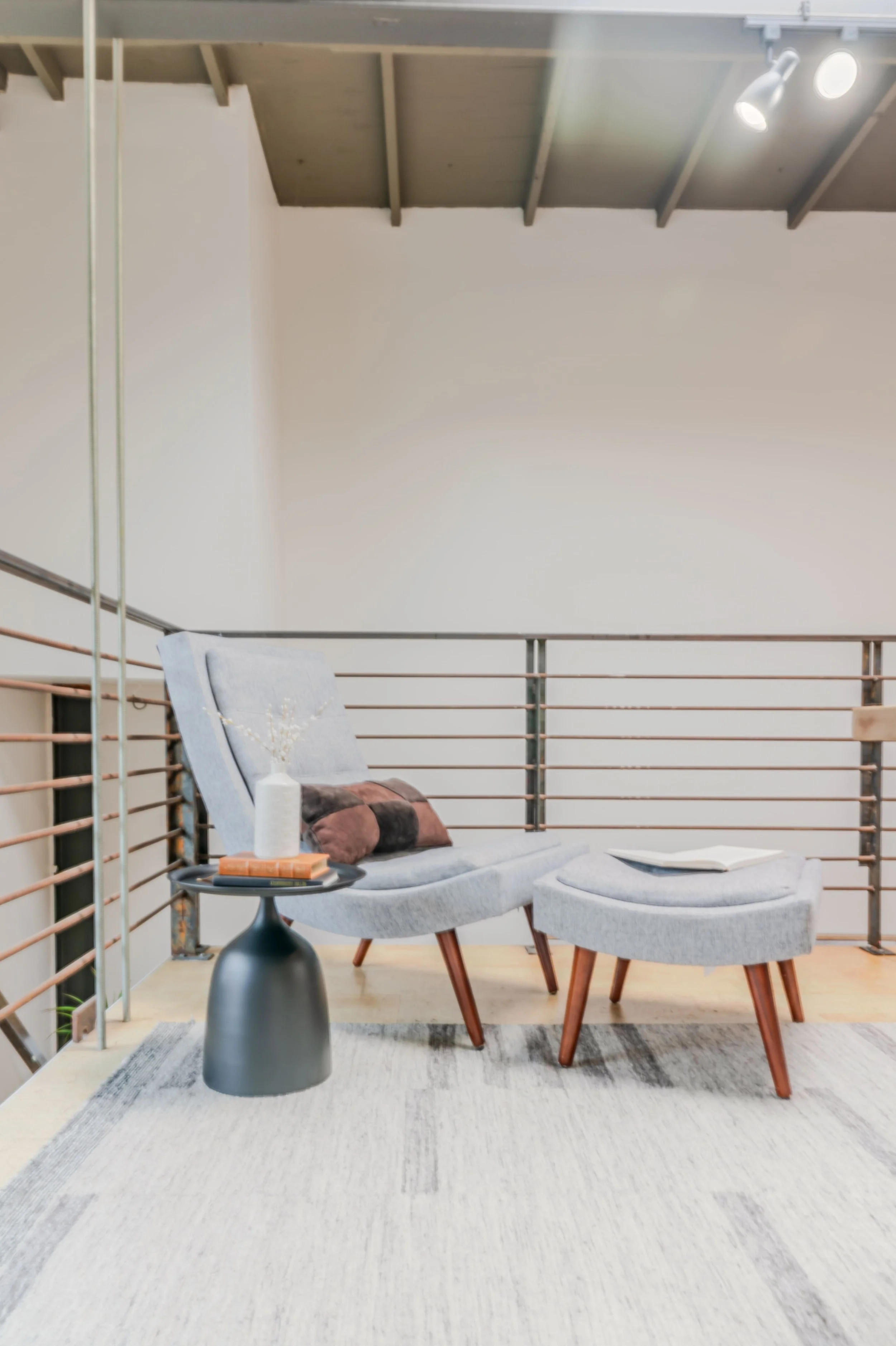 A cozy indoor space with a lounge chair, small side table, and decorative pillows on a rug, featuring wooden ceiling beams and modern lighting.