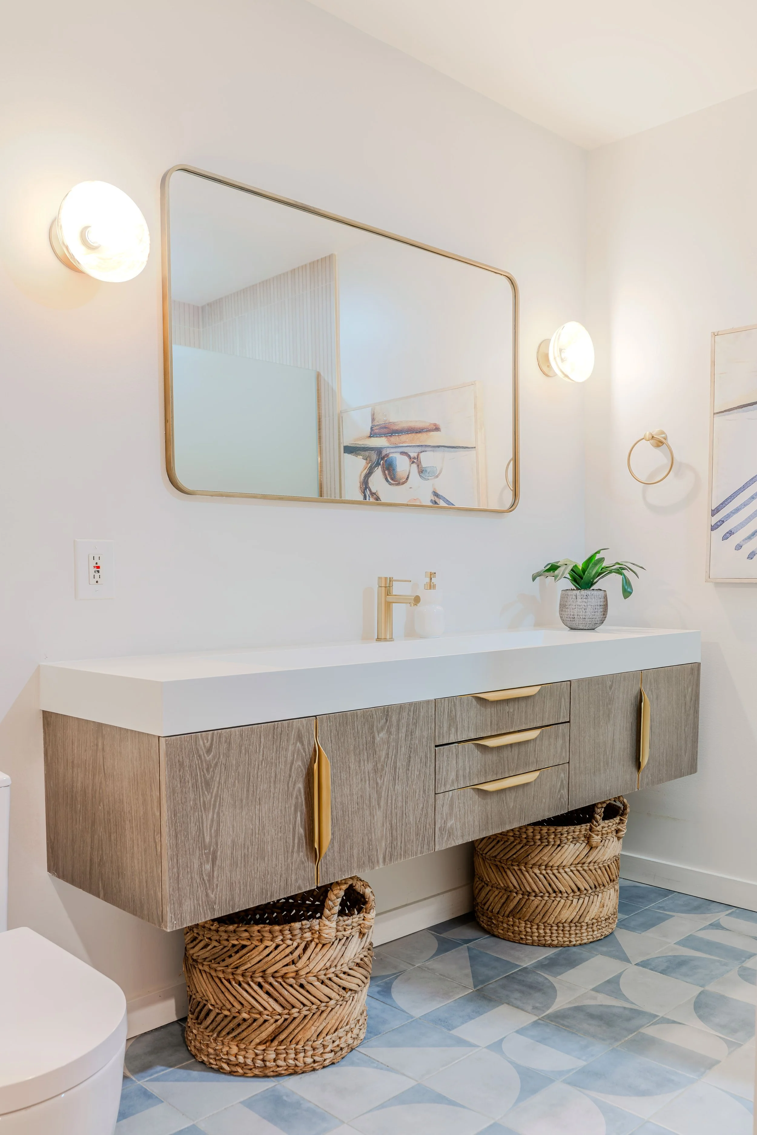 Bathroom vanity with a large mirror, two wicker baskets on the floor, a potted plant, and artwork on the wall.