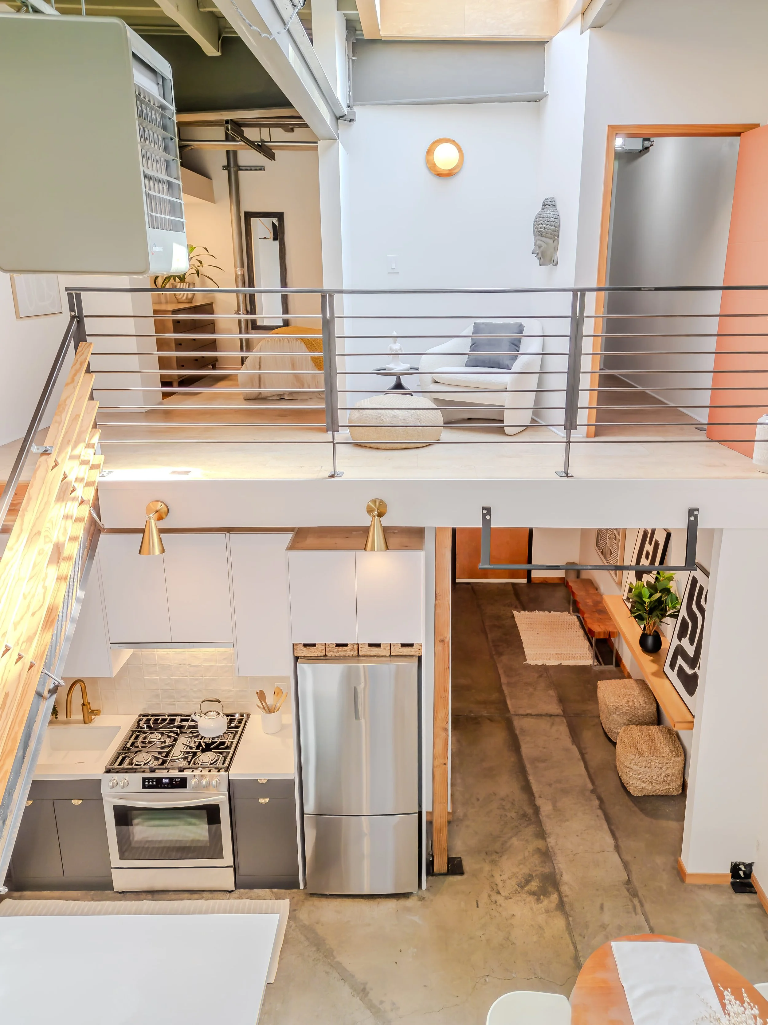 A modern kitchen and living area with stainless steel appliances, including a stove and refrigerator, a staircase with wood steps, a small dining table with a napkin holder, a white armchair with a cushion, abstract artwork, a Buddha head sculpture o