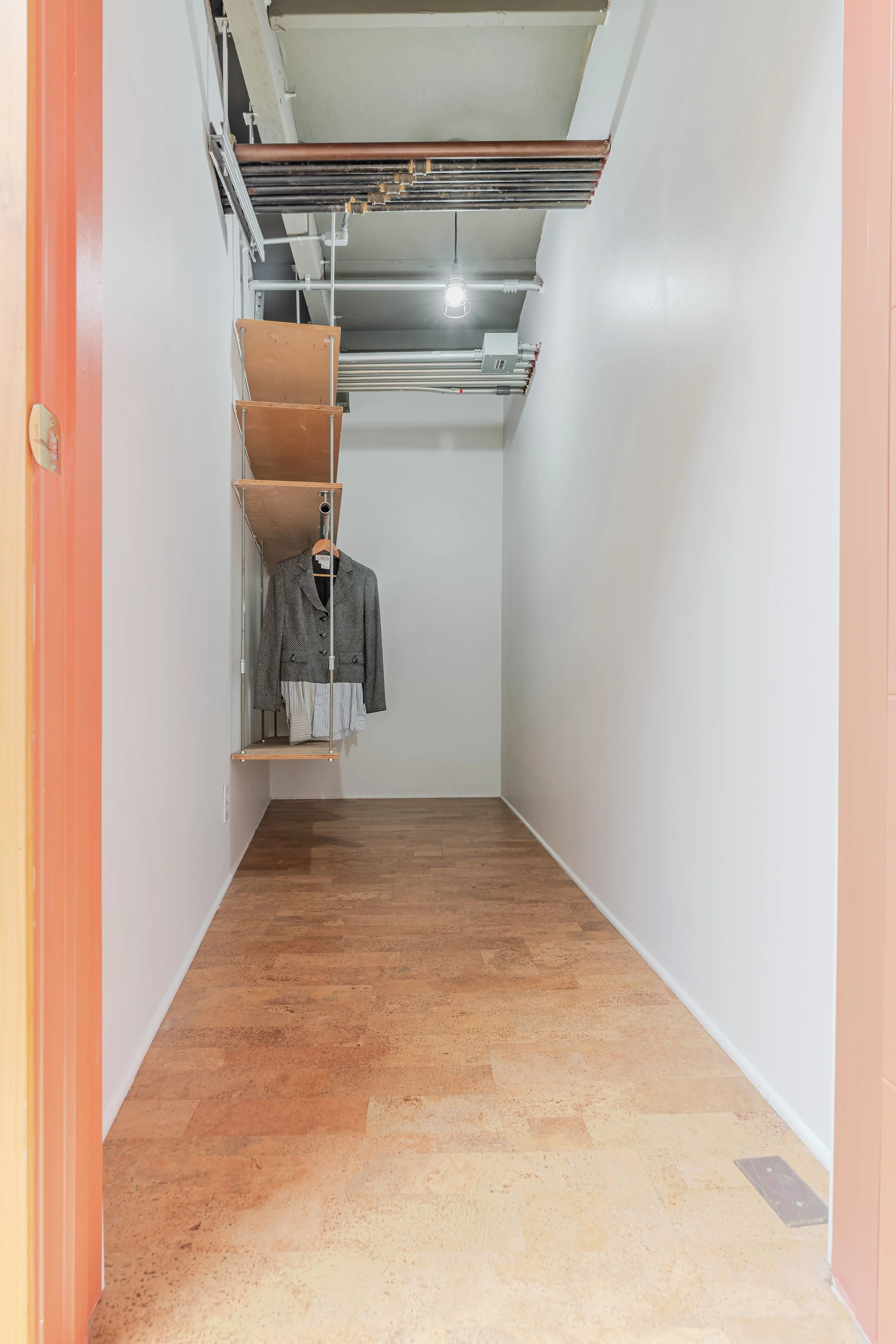 Empty room with white walls and brown wood floor, some plumbing pipes on ceiling, a coat hanging on a metal rod