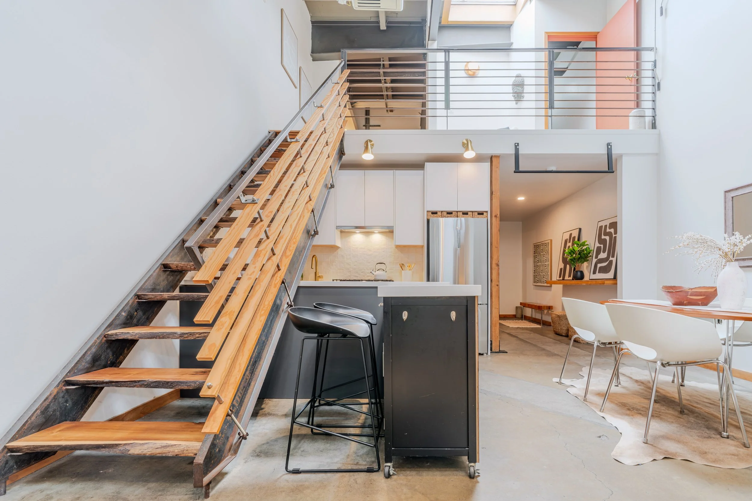 Interior view of a modern, minimalist home with a staircase featuring wooden steps and metal railing, overlooking a dining area with white chairs and a sideboard. Open-concept kitchen in the background with a fridge, white cabinets, and decorative ar