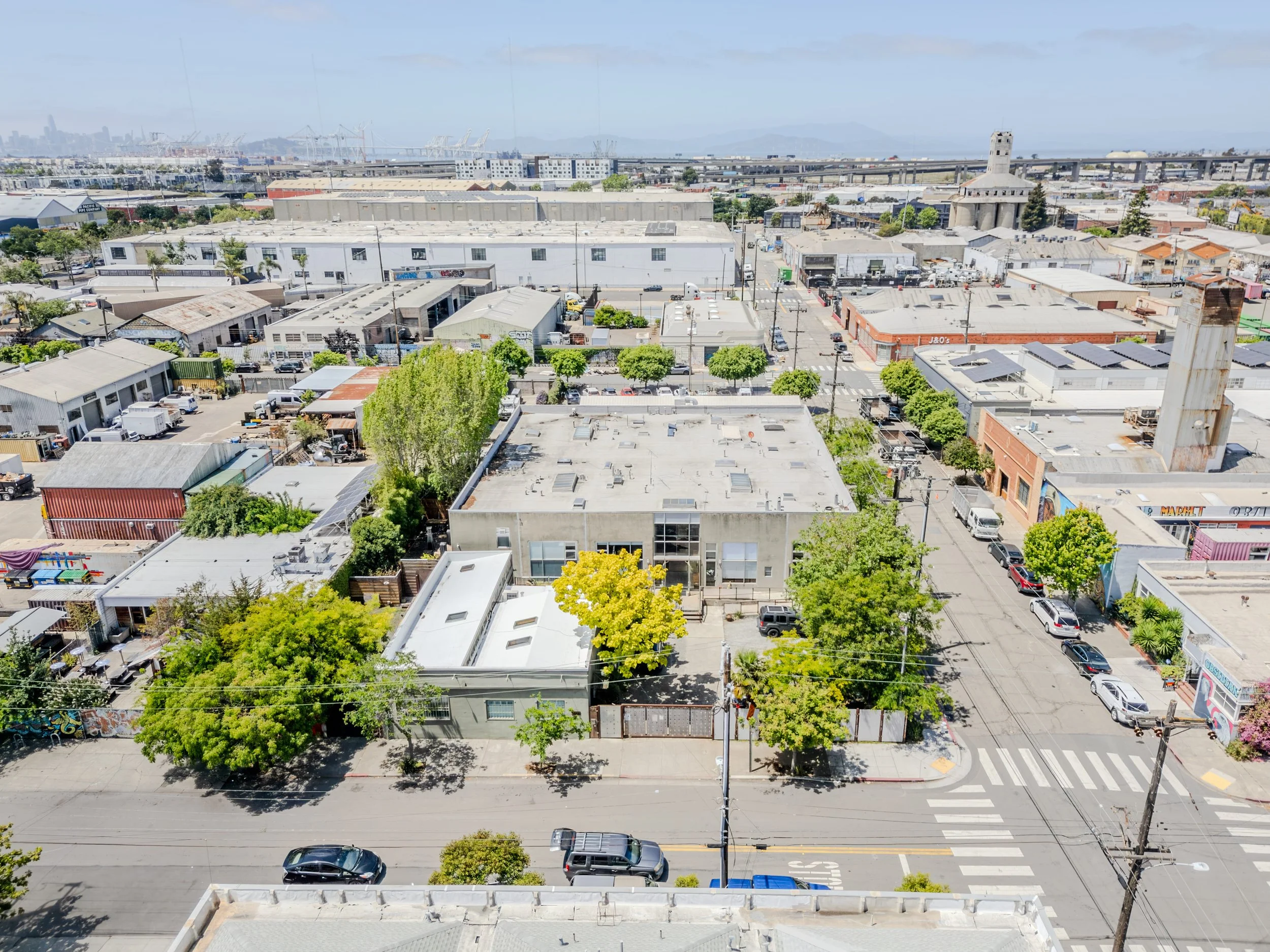 Aerial view of an urban area with various buildings, trees, and parked cars, with a bridge and industrial cranes in the distance under a partly cloudy sky.