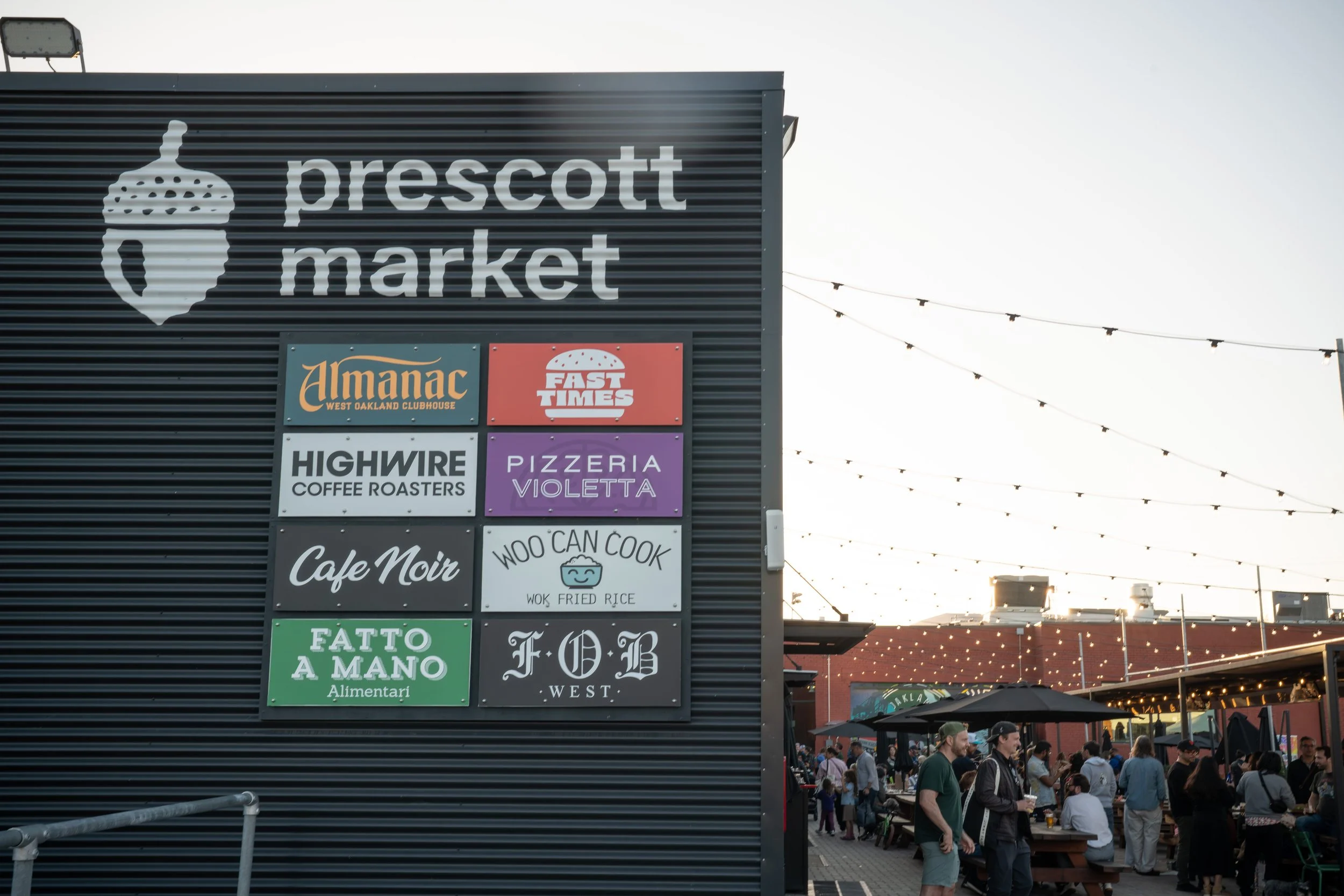 Sign with various business logos at Prescott Market, with people gathered at outdoor seating area during sunset.