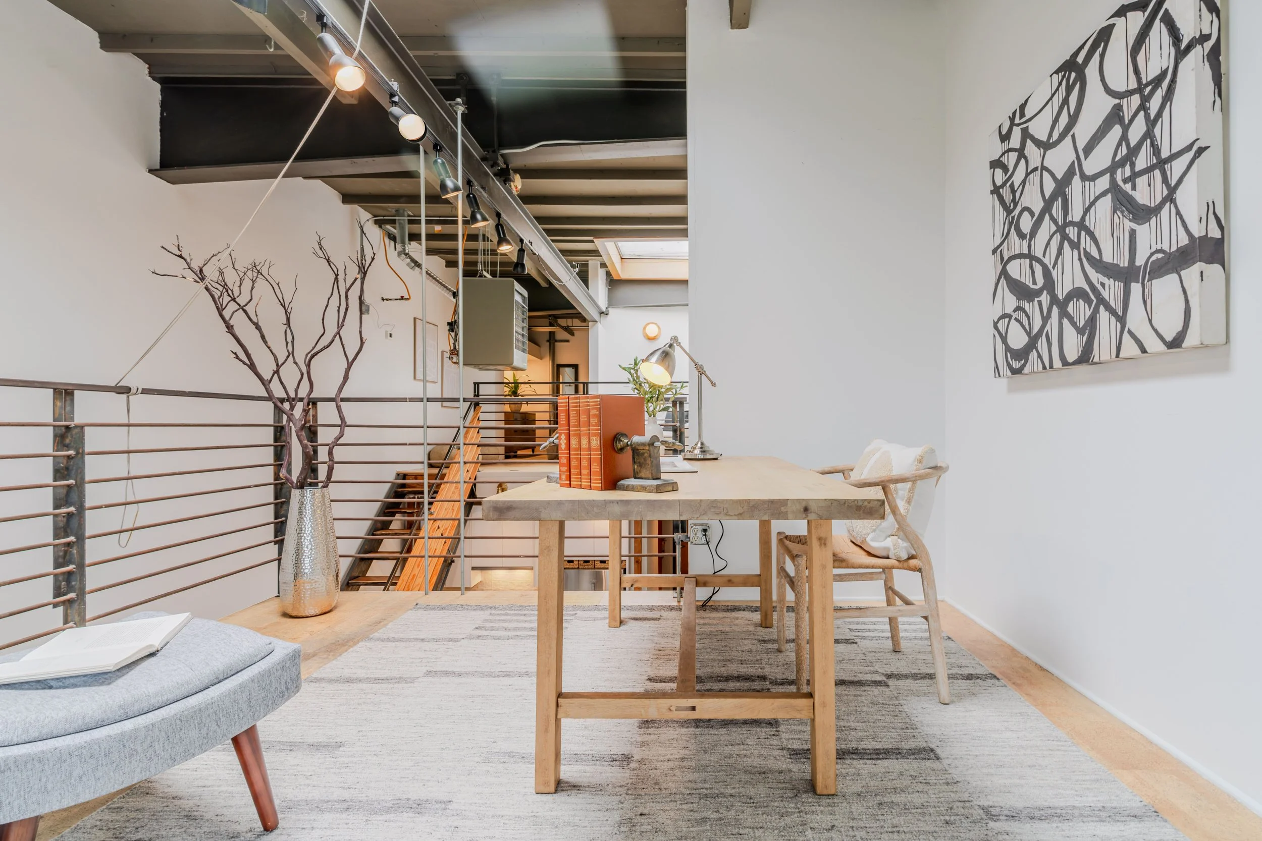 Interior of a modern, minimalist living or working space with a wooden table, wooden chairs, a gray upholstered bench, a tall vase with barren branches, a lamp, books, and wall art with black abstract lines. Exposed ceiling beams and track lighting a
