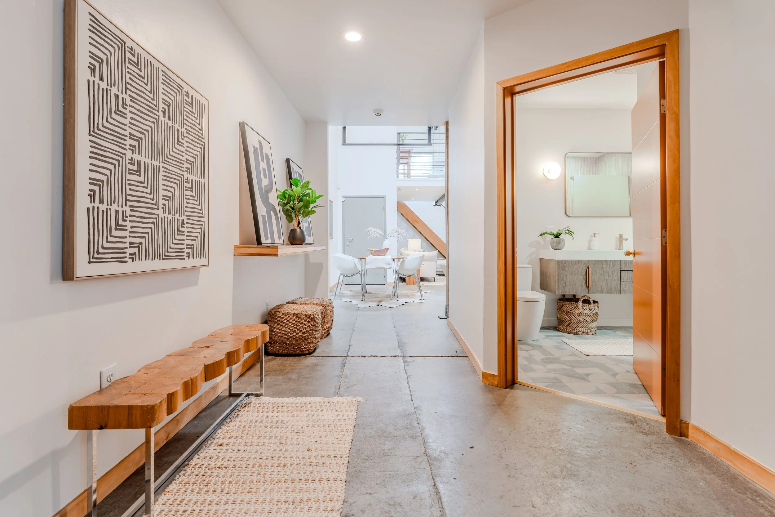 Interior hallway with artwork on white wall, a wooden bench, a plant in a black vase, tiled bathroom with mirror and sink visible through an open door, and a dining area with white chairs and a window at the back.
