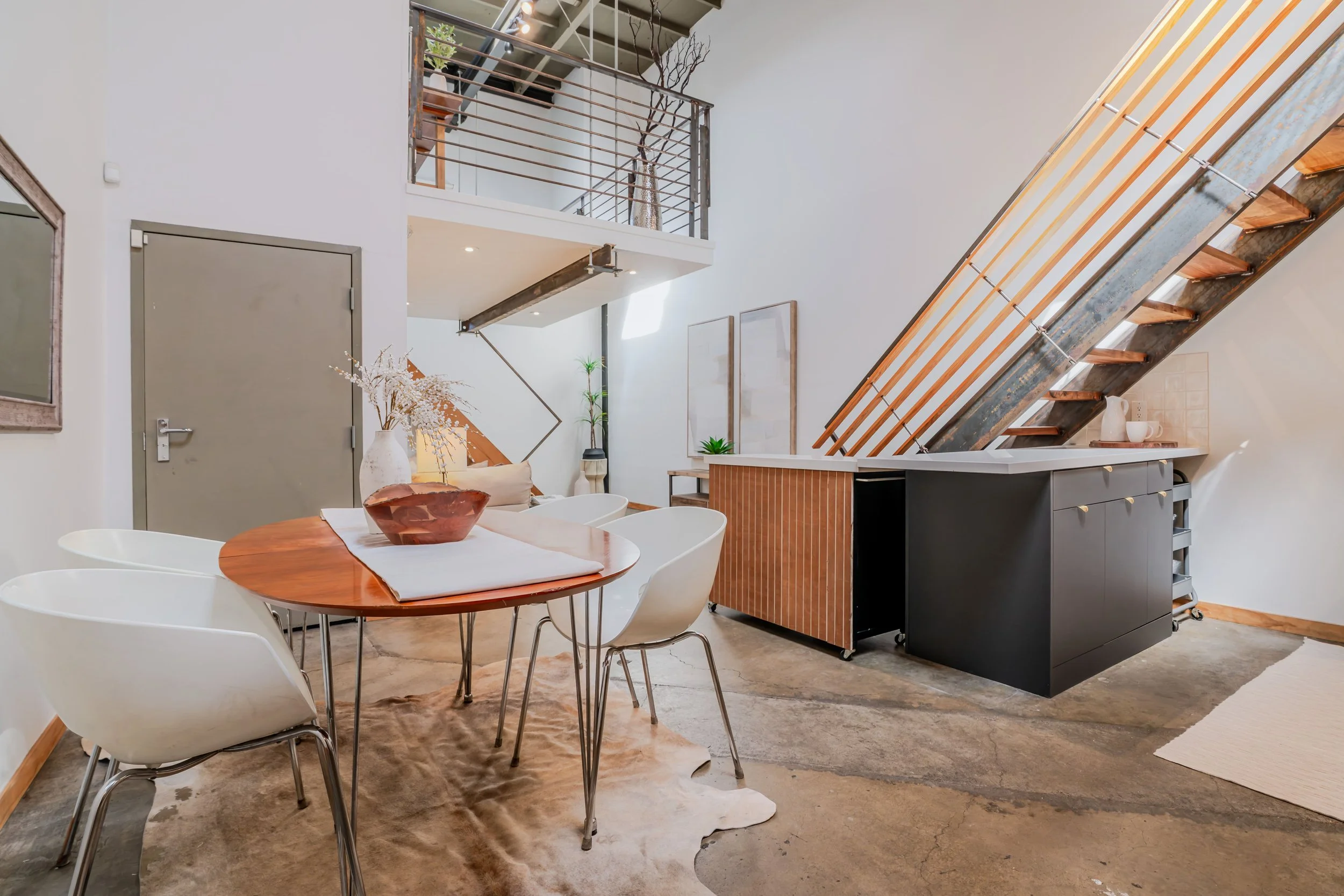 Modern, minimalist dining area with a round wooden table surrounded by white chairs, a cowhide rug on a concrete floor, and a kitchen with black cabinetry, wooden accents, and an industrial style staircase leading to an upper level, with decorative plants and artwork on the walls.