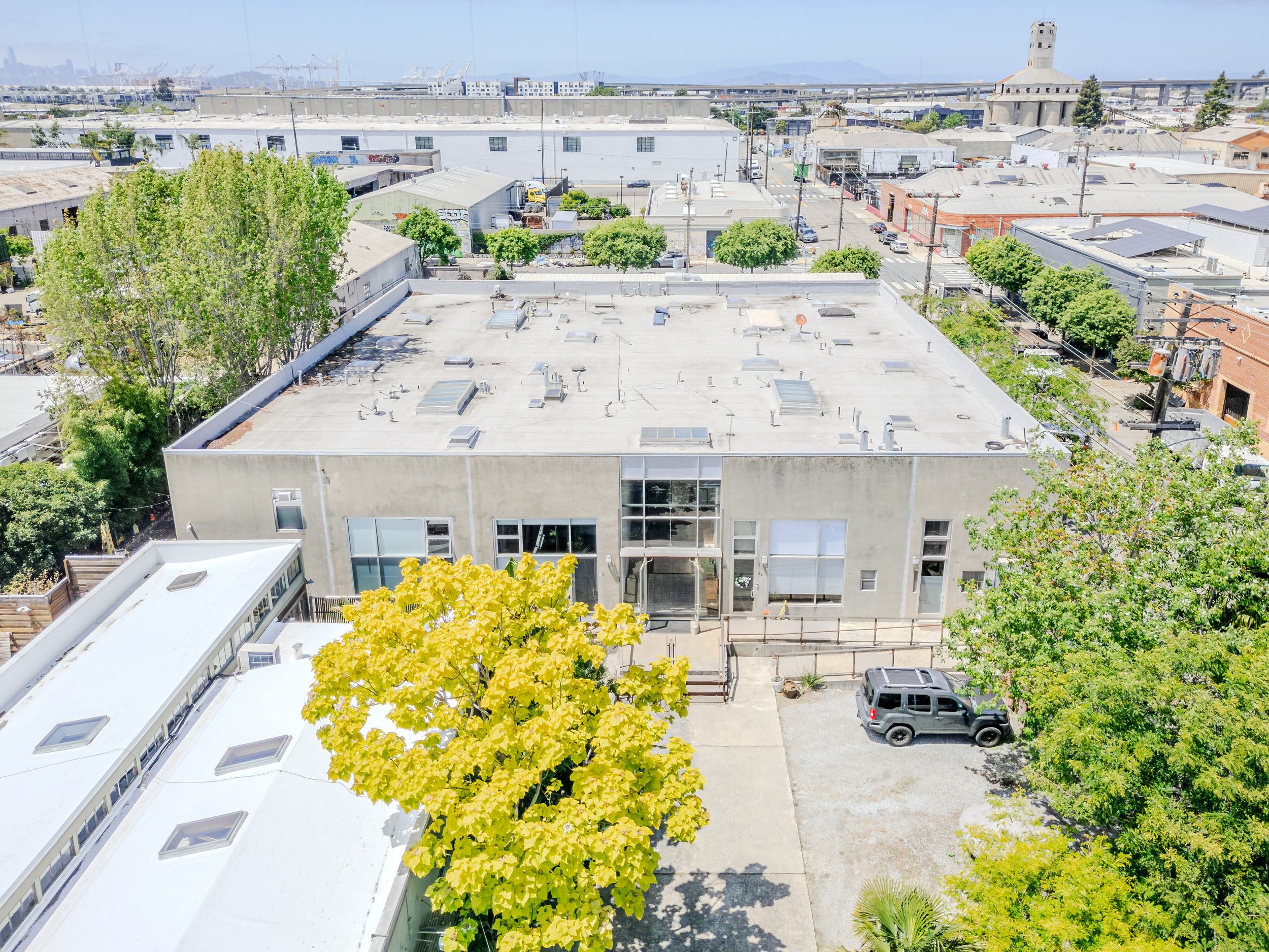 An urban rooftop view with green trees, parked cars, surrounding buildings, and a distant industrial area under a clear sky.
