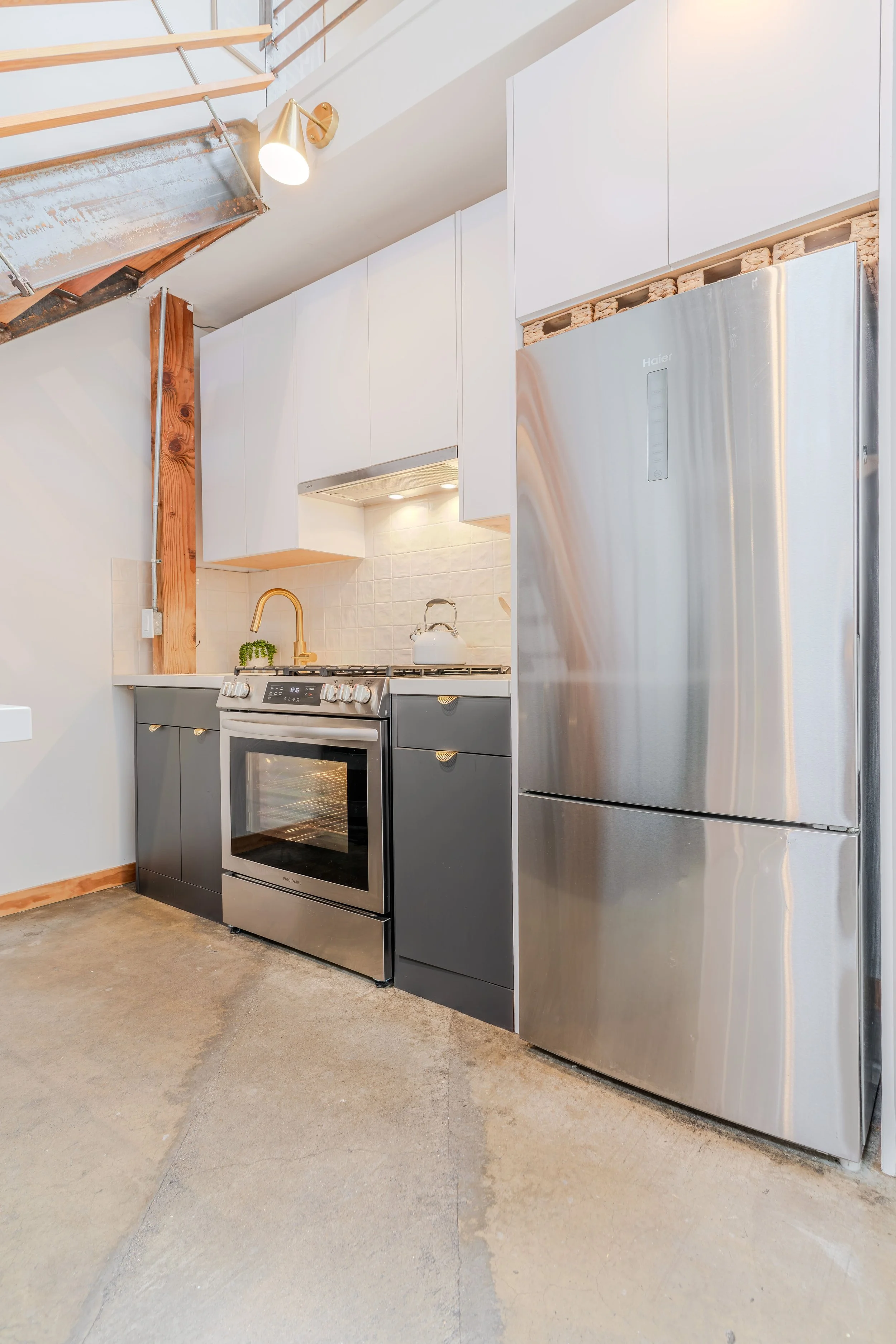 Modern kitchen with white cabinets, stainless steel refrigerator, stove, and a gold sink faucet. The ceiling has a wooden beam, and the floor appears unfinished concrete.